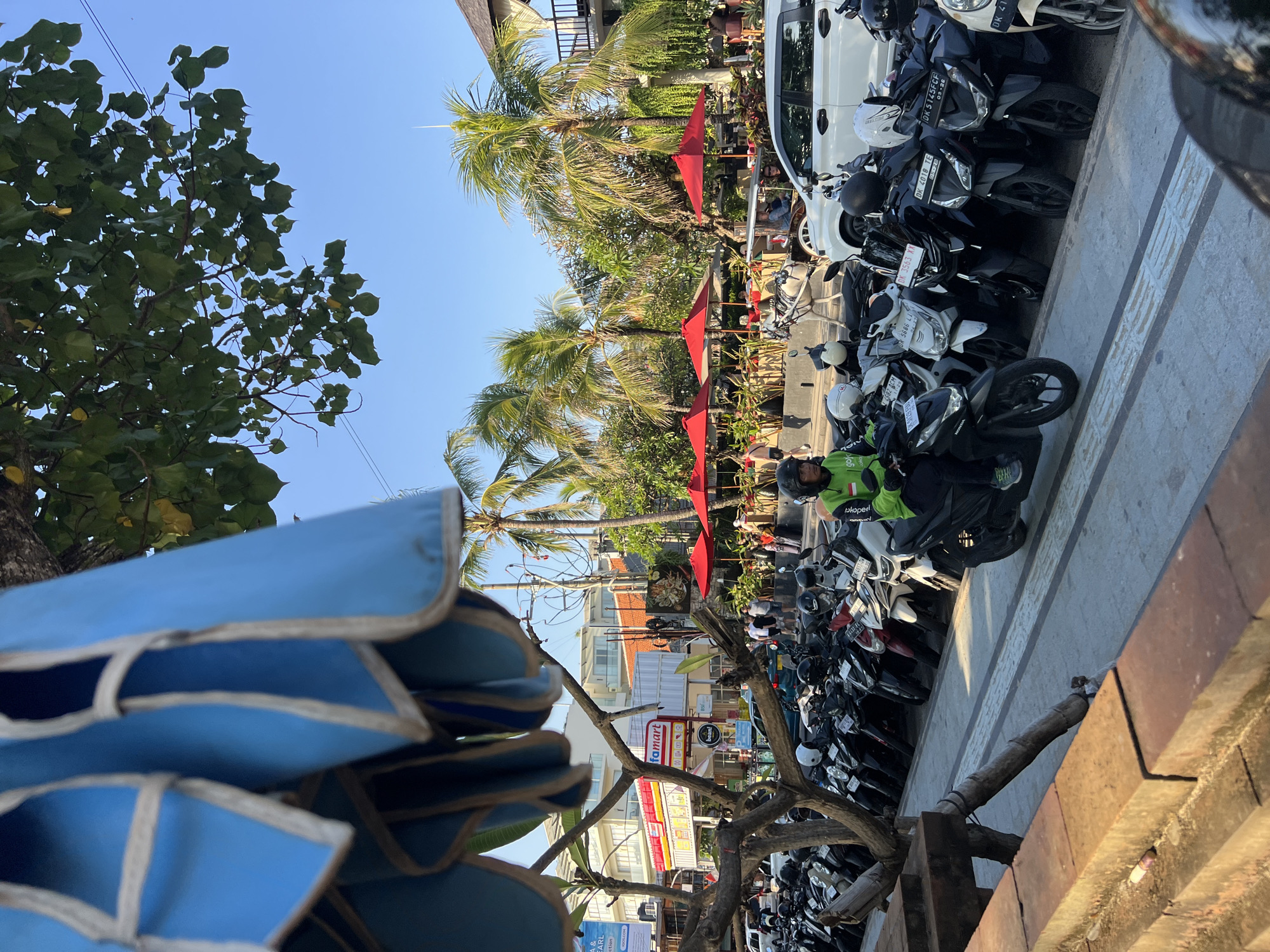 The image depicts a street scene in Kuta, Indonesia, likely during daylight hours under a clear blue sky. In the foreground, a line of parked motorcycles stretches along a paved walkway. A person wearing a bright green jacket with a Gojek logo is riding one of the motorcycles. Beyond the motorcycles, there are several red umbrellas set up, indicating an outdoor seating area for a cafe or restaurant. Palm trees are visible in the background, and there is a building with a "Famart" sign, suggesting a commercial area. A white car is parked near the outdoor seating area. The overall impression is of a bustling tropical town.