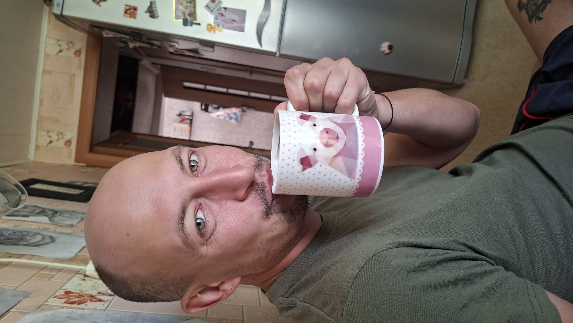 A bald man with light-colored eyes and a goatee holds a white mug with a pink pig design to his lips. He is wearing a green t-shirt. The background appears to be a kitchen with patterned wallpaper, a doorway, and a refrigerator. The image is cropped to focus on the man and the mug. The location is the city of Myadzyel, Belarus.