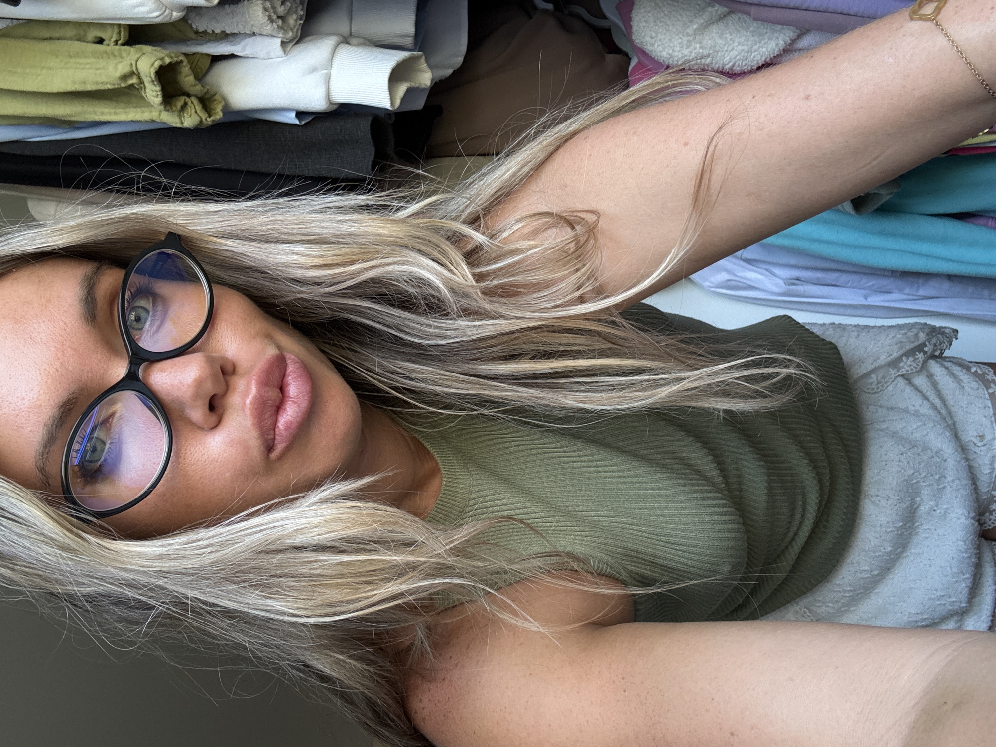 The image is a close-up, slightly angled selfie of a woman with blonde hair and glasses. She is wearing a green ribbed tank top and white lace shorts. Her lips are pursed. In the background, clothing is visible hanging in what appears to be a closet. The scene is indoors with natural light coming from the left. There is no identifiable information to place the location in Votkinsk, Russia.