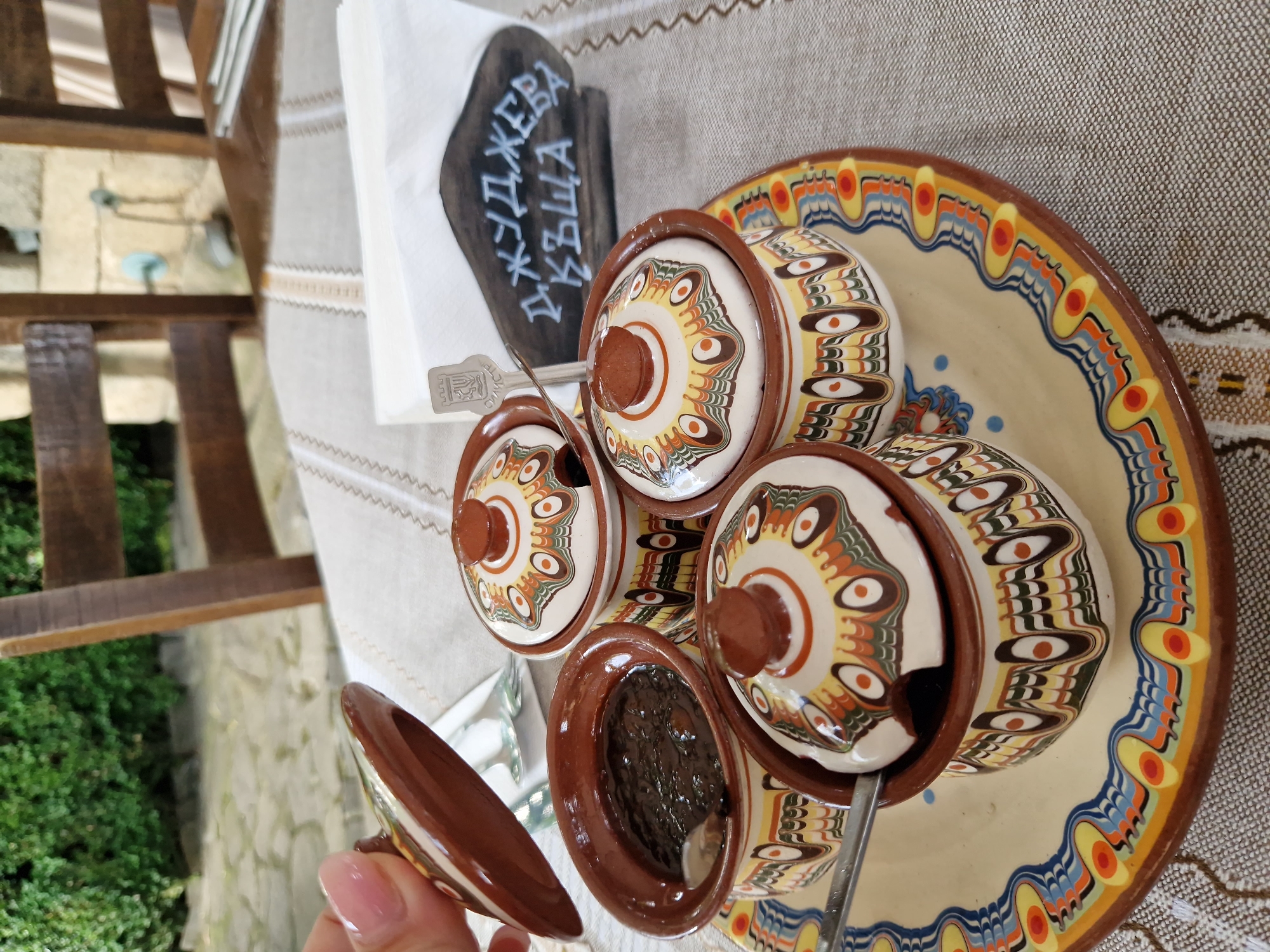 A hand with a pink, glossy fingernail holds the lid of a small, decorative ceramic pot. The pot is part of a set of similar containers arranged on a larger ceramic plate. These pots and the plate are adorned with a traditional folk art design featuring concentric circles, wavy lines, and eye-like motifs in earthy tones of brown, yellow, orange, and green. One of the pots, open, contains a dark, chunky substance, likely a jam or preserve, with a small spoon resting inside it. Another pot has a spoon partially inserted into it. In the background, a wooden napkin holder with an embossed emblem and white lettering in Bulgarian, reading "ДЖУДЖЕВА КЪЩА" (Dzhudzheva Kashta - Dwarf House), sits on the table next to a stack of white napkins. The table is covered with a textured, neutral-colored tablecloth. Wooden chairs and a stone wall are visible in the blurred background, suggesting an outdoor seating area. The overall scene depicts a close-up of a traditional Bulgarian culinary offering, likely in a restaurant or cafe setting in Panagyurishte, Bulgaria, given the distinctive pottery style and the name on the napkin holder which is associated with a themed attraction there.