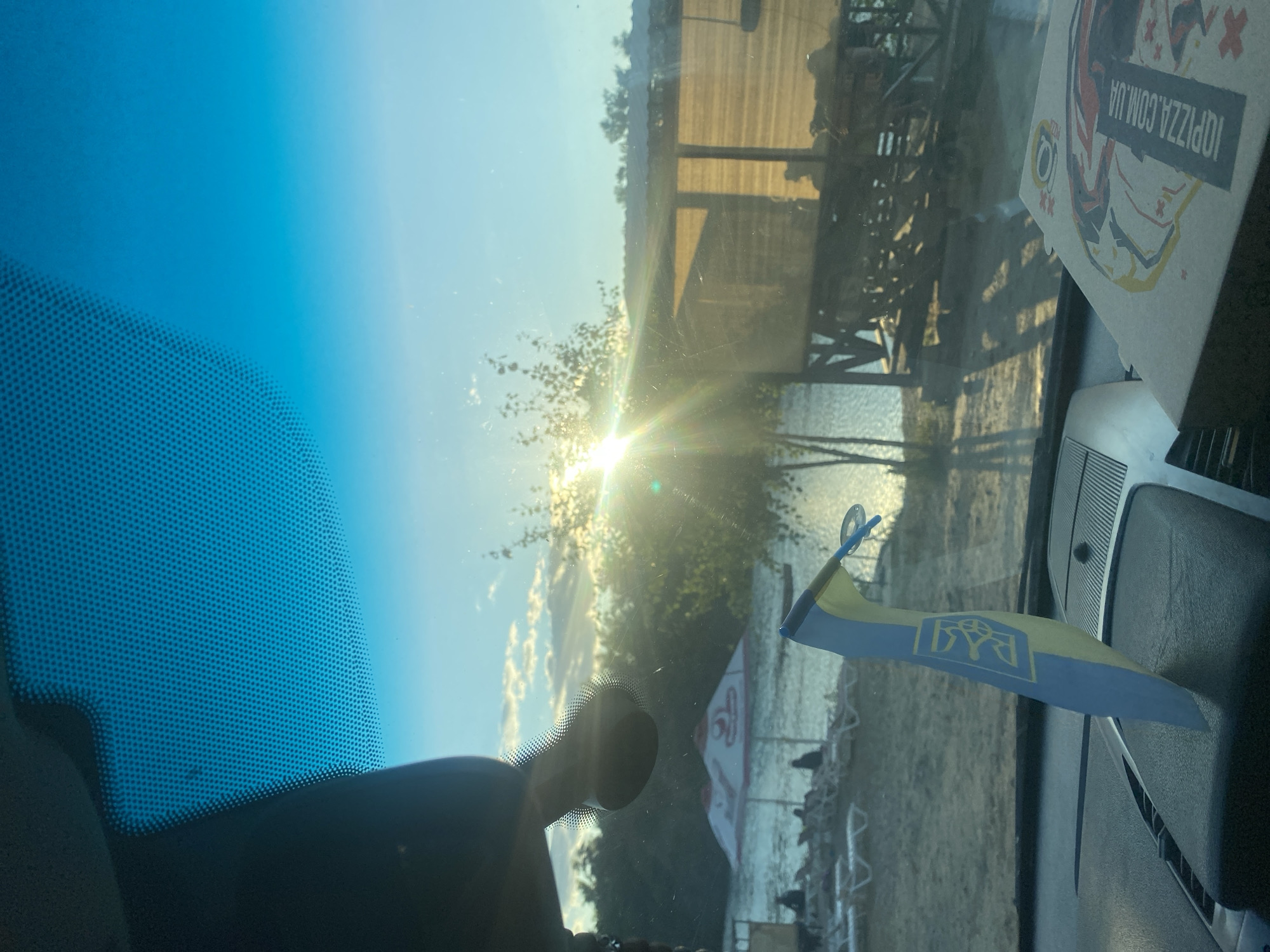 The image is taken from inside a vehicle, looking through the windshield. The sun is setting over a body of water, casting a bright glare. In the foreground, on the dashboard, is a pizza box with the logo "IQ PIZZA.COM.UA". A Ukrainian flag is also visible on the dashboard, positioned in front of the windshield. Beyond the windshield, a beach scene is depicted with beach chairs and umbrellas. A wooden structure, possibly a cafe or restaurant, is situated on the right side of the frame, adjacent to the water. The background shows trees and a clear sky. The location is identified as the Dnipro River embankment in Dnipro, Ukraine.