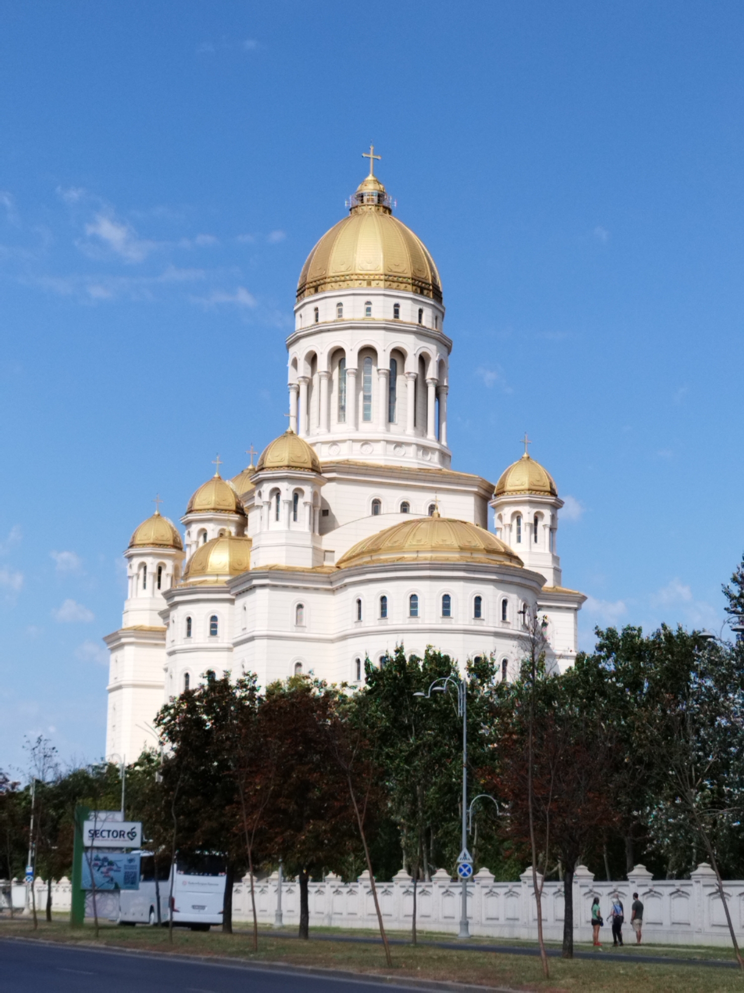 A large, white cathedral with golden domes dominates the center of the image, under a clear blue sky with a few wispy clouds. The cathedral features classical architecture with columns and arched windows. In the foreground, a white bus is parked to the left of a fence. Several trees, some with dark green foliage and others with reddish-brown leaves, are positioned between the cathedral and the street. A tall lamp post with a curved arm stands near the fence. A white sign with the word "SECTOR" and a logo is visible in front of the bus. Three people are standing on a sidewalk to the right of the fence, facing the cathedral. The scene appears to be in Bucharest, Romania.