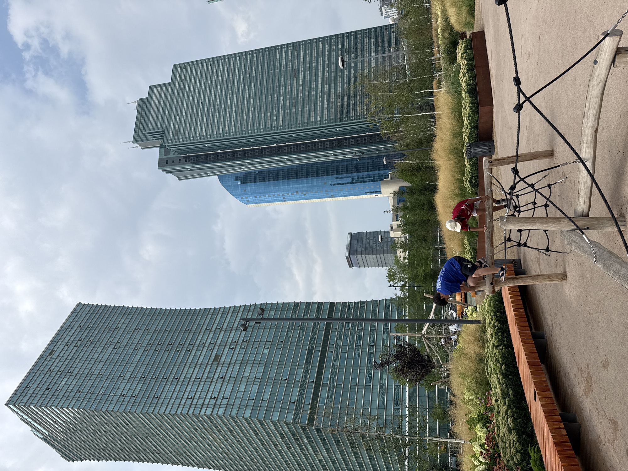 This is an exterior, eye-level, full shot of the Nur Alem pavilion, part of the EXPO 2017 complex in Astana, Kazakhstan, on a cloudy day. The futuristic, spherical building, clad in glass and metal, dominates the center of the image. In the foreground, two young boys are engaged in climbing on a wooden play structure. The boy on the left, wearing a blue t-shirt and black shorts, is positioned on top of a horizontal beam. The boy on the right, in a red t-shirt and white cap, is also climbing on the structure. Surrounding the play area is a landscaped park with various types of plants, including tall grasses, bushes, and trees. A curved wooden bench runs along the left side of the frame. In the background, beyond the pavilion, are other modern skyscrapers, characteristic of Astana's cityscape, including a distinctive blue glass building and several tall, slender towers with glass facades. The sky is overcast with scattered clouds.