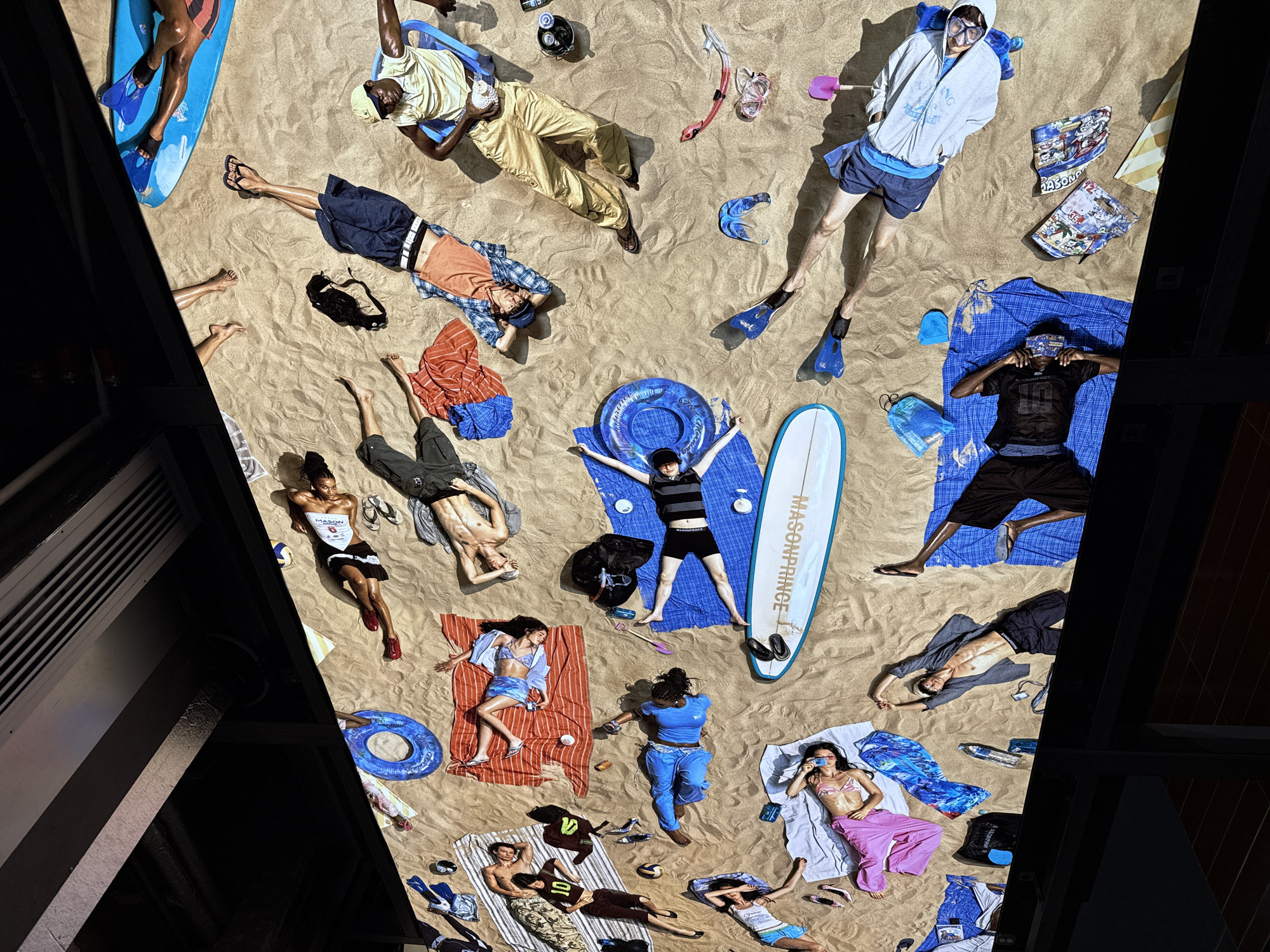 An overhead shot reveals a striking art installation or staged scene, simulating a lively beach environment, likely within an exhibition space in Tianhe District, China. Approximately 20 to 25 young adults are depicted lying or sitting on artificial sand, surrounded by a multitude of beach-related items.
The individuals are dressed in diverse beach attire, ranging from swim shorts, bikinis, and t-shirts to casual layered clothing. Some are equipped with diving masks, snorkels, and flippers, suggesting a playful, leisure-focused atmosphere. The sandy surface is meticulously detailed, showing textures and subtle footprints, contributing to the realism of the faux beach.
Scattered across the "sand" are numerous props: two surfboards (one prominently branded "MASONPRINCE"), a boogie board, bright blue inflatable rings, colorful beach towels, beach chairs, sunglasses, water bottles, various bags (another bearing "MASONPRINCE"), sandals, a volleyball, and a small pink shovel.
The scene is illuminated by bright, consistent overhead lighting, casting strong, distinct shadows that mimic a sunny day. Dark, angular architectural structures flank the 'beach' on the left and right, clearly delineating the edges of the installation and underscoring its indoor, constructed nature. The people appear relaxed and calm, engaging in various states of repose, from sunbathing to contemplative lounging. Visible text includes "MASONPRINCE" on a surfboard and a bag, "MASON" on a white top, and "10" on a dark top.