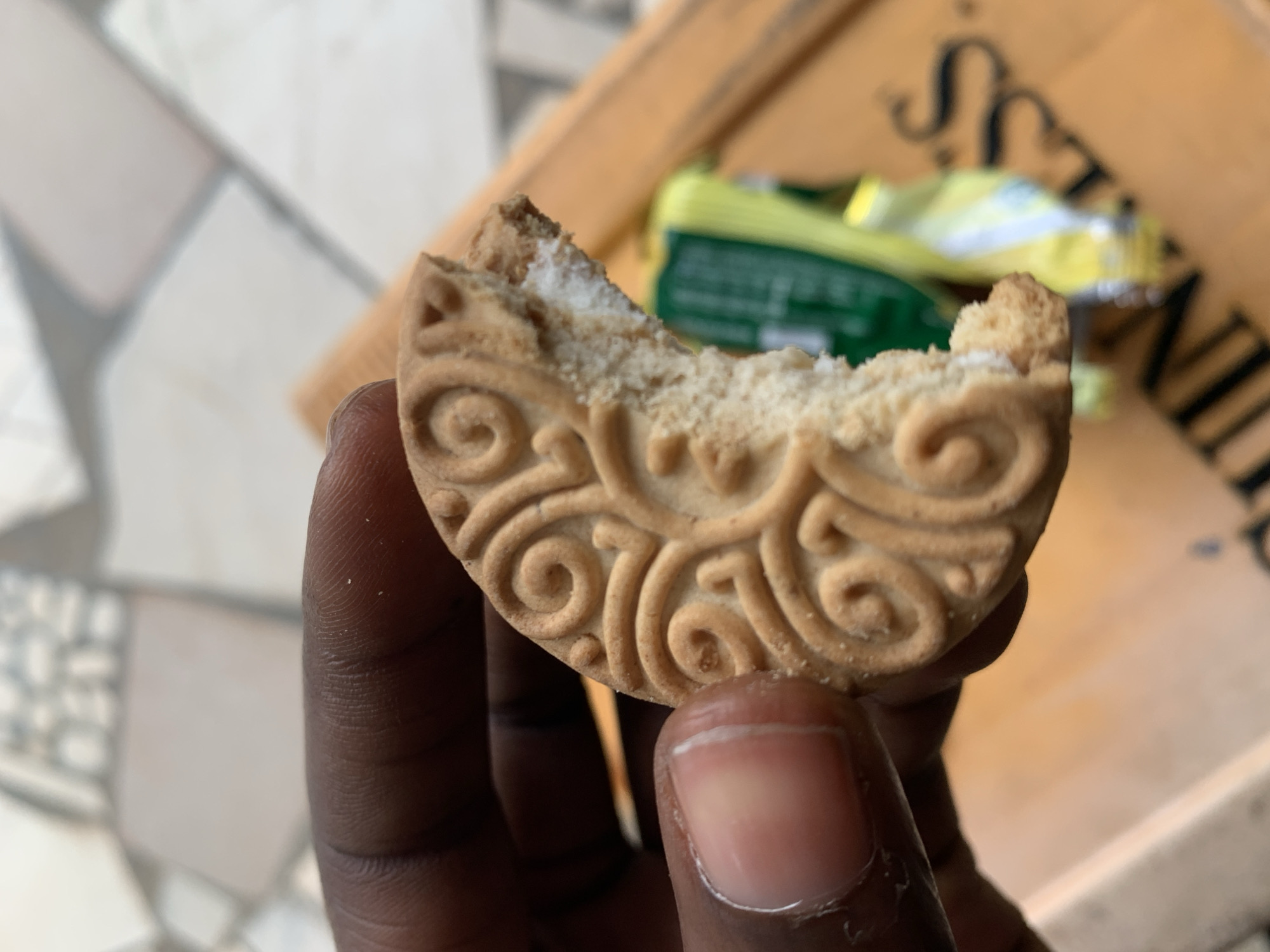 A partially eaten biscuit, decorated with swirls, is held in a dark-skinned hand with short fingernails. In the blurred background, a wooden surface bearing text (illegible) and the partially visible packaging of the biscuit are present. The surface beneath the hand appears to be tiled. No explicit evidence suggests this image was taken in New Stadium, Nigeria. No people are depicted other than the implied presence of the hand's owner.