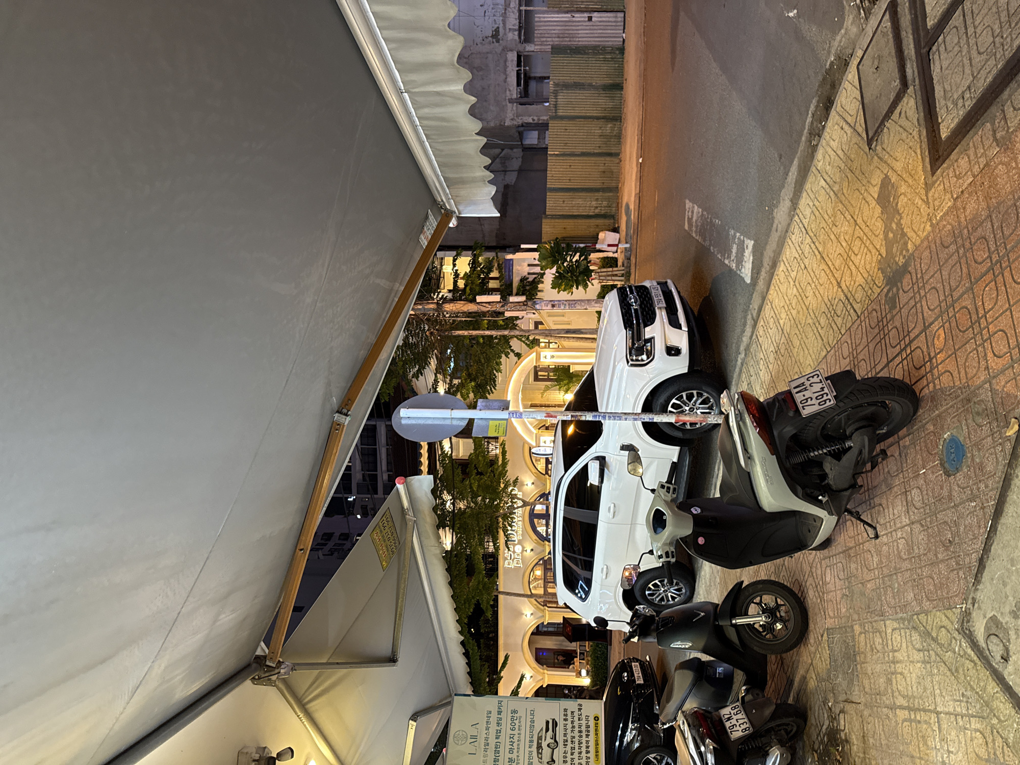 The image depicts a street scene in Nha Trang, Vietnam, likely in the evening or at night, given the artificial lighting. An awning extends over a sidewalk area where several motorcycles are parked. A white SUV is parked on the street in front of a building with illuminated arches. The building appears to be a restaurant or cafe. A sign in Korean is visible on a stand on the left side of the image. The street is paved with patterned tiles, and a road with white lane markings is visible to the right. The general atmosphere is that of an urban commercial district.