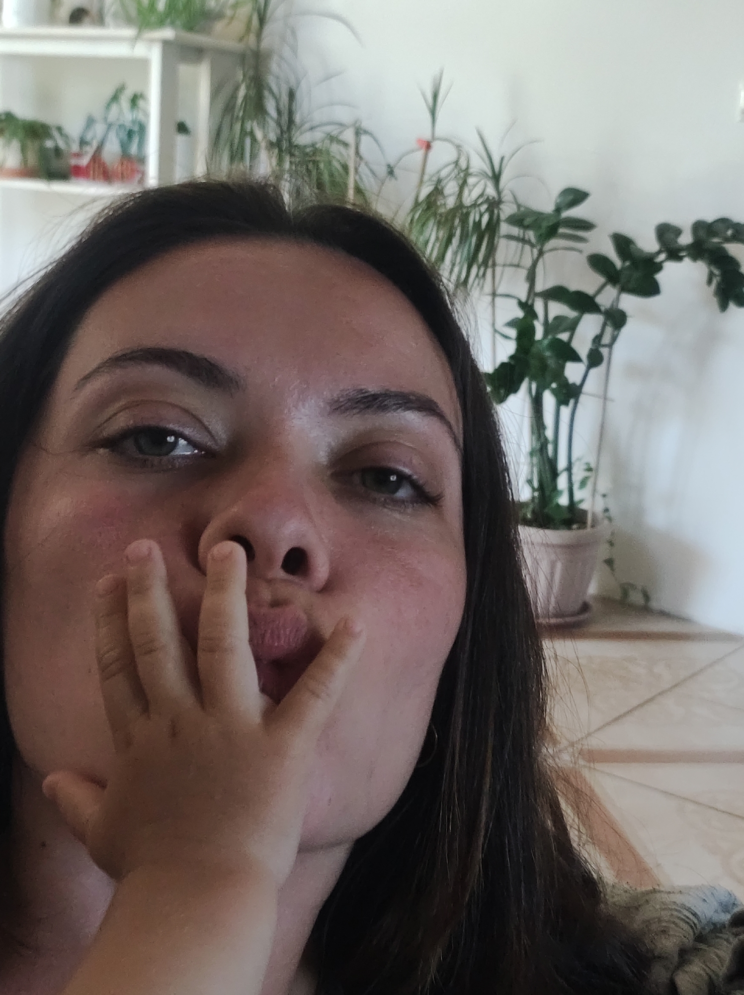 A close-up selfie shows a woman with dark hair and brown eyes, her face angled towards the camera as she puckers her lips. A child's hand is covering part of her mouth. In the background, a white wall is adorned with several potted plants. The floor is tiled in a light brown and cream pattern.