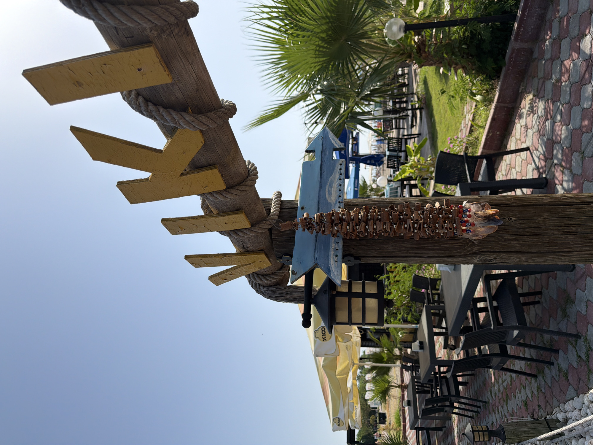 A low-angle medium shot captures a wooden post adorned with a rustic hanging decoration and a blue arrow-shaped sign, with the letters "VIKI" painted in yellow on a wooden beam overhead. The scene is set outdoors in Kemer, Turkey, suggesting a coastal or beachside establishment. Tables and chairs are arranged on a patterned paver patio, with umbrellas and palm trees indicating a warm, sunny climate. In the background, a beach and the sea are visible under a clear blue sky. The overall ambiance is relaxed and inviting, hinting at a casual dining or relaxation area.