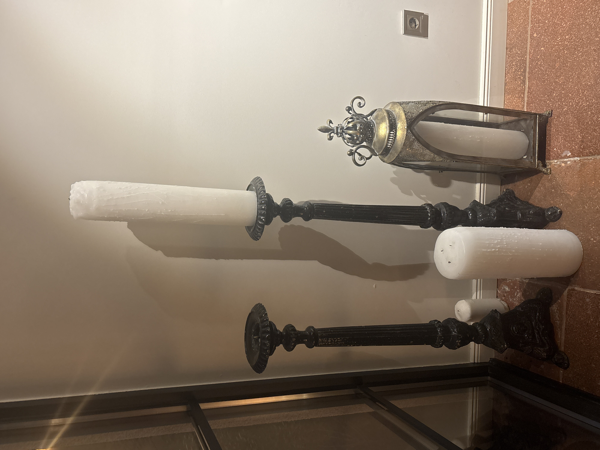 A vertical, medium shot shows four white pillar candles in ornate, black metal candleholders and an ornate gold and silver metal lantern on a white surface, with a red surface to the right. The largest pillar candle is set in a candleholder near the top left corner of the image. A metal ring is set around the base of the candle. To the right is a second pillar candle in an ornate lantern with a decorative top. To the right of the first candle and below the lantern is a third, shorter pillar candle, resting without a candleholder. The smallest candle is set in a candleholder on the bottom right corner. A small light switch can be seen on the white wall, and the top of a black frame is seen along the bottom of the image.