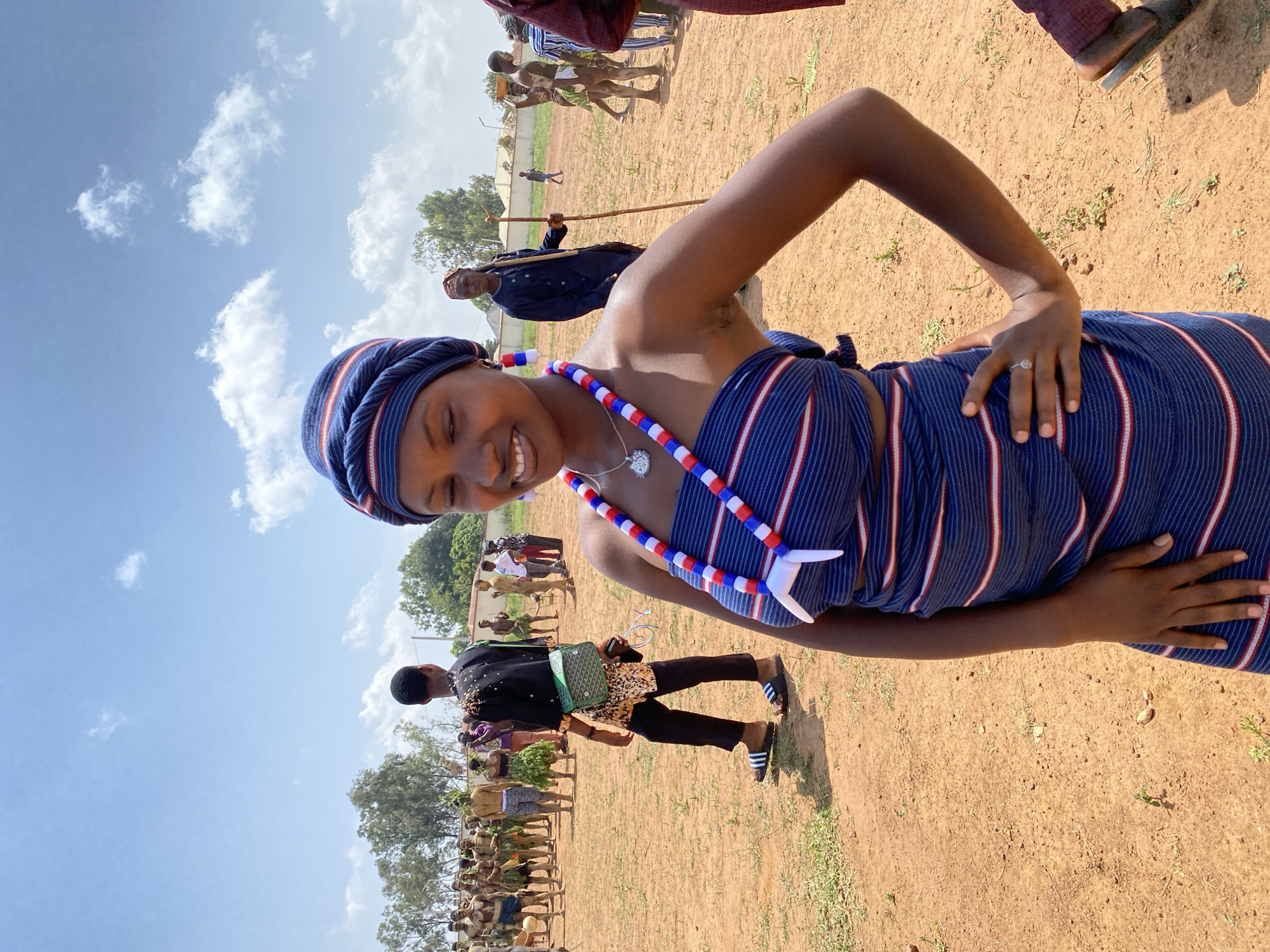 The image depicts a woman in traditional attire, including a blue and white striped cloth wrapped around her body and a matching headwrap. She is also wearing a red, white, and blue beaded necklace with a white pendant. She is smiling and has one hand on her hip. The location appears to be an outdoor setting with a reddish-brown dirt ground and a group of people in the background, some also in traditional clothing. The sky is visible with light clouds. There is no explicit indication of Durimi, Nigeria.