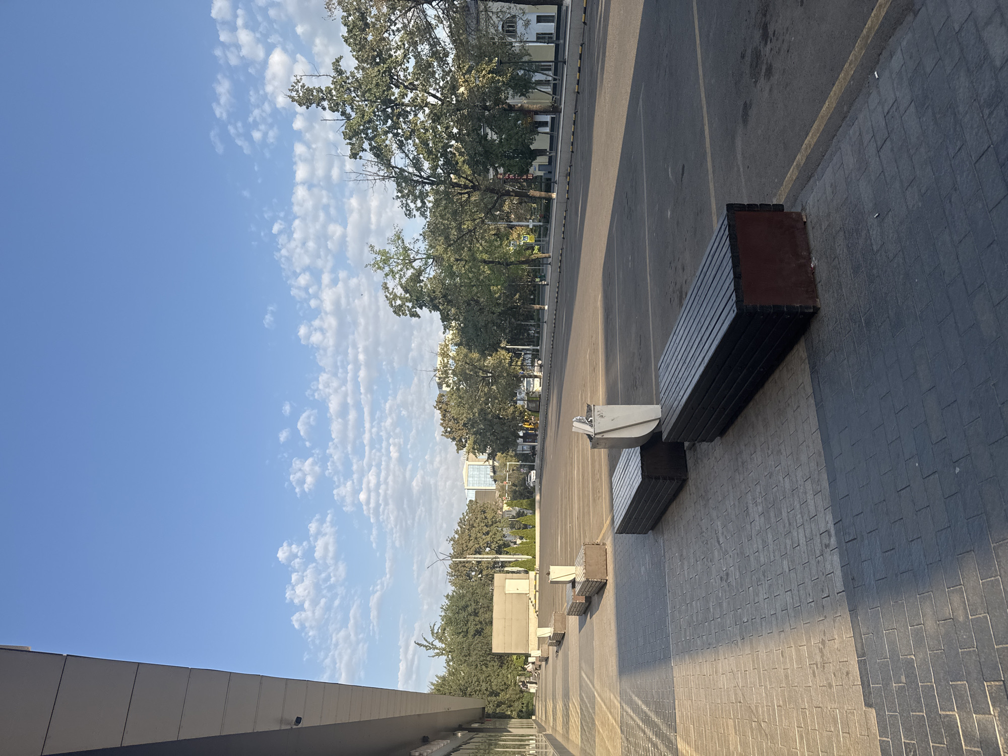 The image captures an outdoor scene in Bishkek City, Kyrgyzstan, likely a public space adjacent to a building and a parking lot. The time of day appears to be morning or late afternoon, given the long shadows cast by the building and the lighting conditions.
In the foreground, a paved walkway made of rectangular brick-like stones is visible. Sunlight illuminates a portion of this walkway, creating distinct shadow patterns from an unseen structure. Several dark-colored, slatted benches with red bases are placed along the walkway. One bench, in the immediate foreground, is largely in shadow. Further back, another bench is illuminated. A metal trash receptacle is positioned behind this second bench.
The walkway leads to a vast asphalt parking lot, marked with yellow lines. The parking lot is mostly empty, with only a few distant vehicles visible. Trees line the far side of the parking lot, with their branches extending into the frame. Behind the trees, buildings of varying heights and architectural styles are present, including a modern glass-fronted building and older, more traditional structures.
The sky above is a clear blue with scattered white, cumulus clouds that are elongated and stretched out. The overall impression is one of urban emptiness and tranquility under a bright sky. A few people are visible in the distance walking along the walkway near the building.