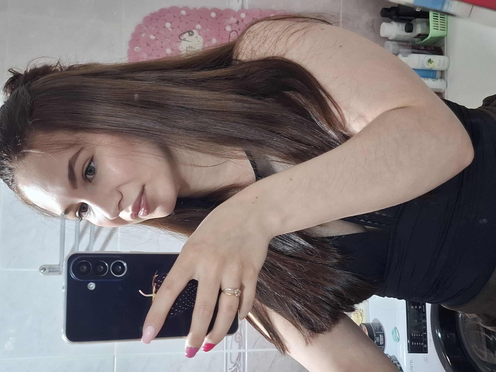 A young woman is taking a selfie in a bathroom. She has long brown hair and is wearing a black top. She is holding a dark-colored smartphone with multiple camera lenses. She has a ring on her finger and her nails are painted in different shades of pink. The background includes a tiled wall, a pink bath mat, shelves with various items, and a washing machine. The location is likely a private residence in the city of Chaikovsky, Russia.