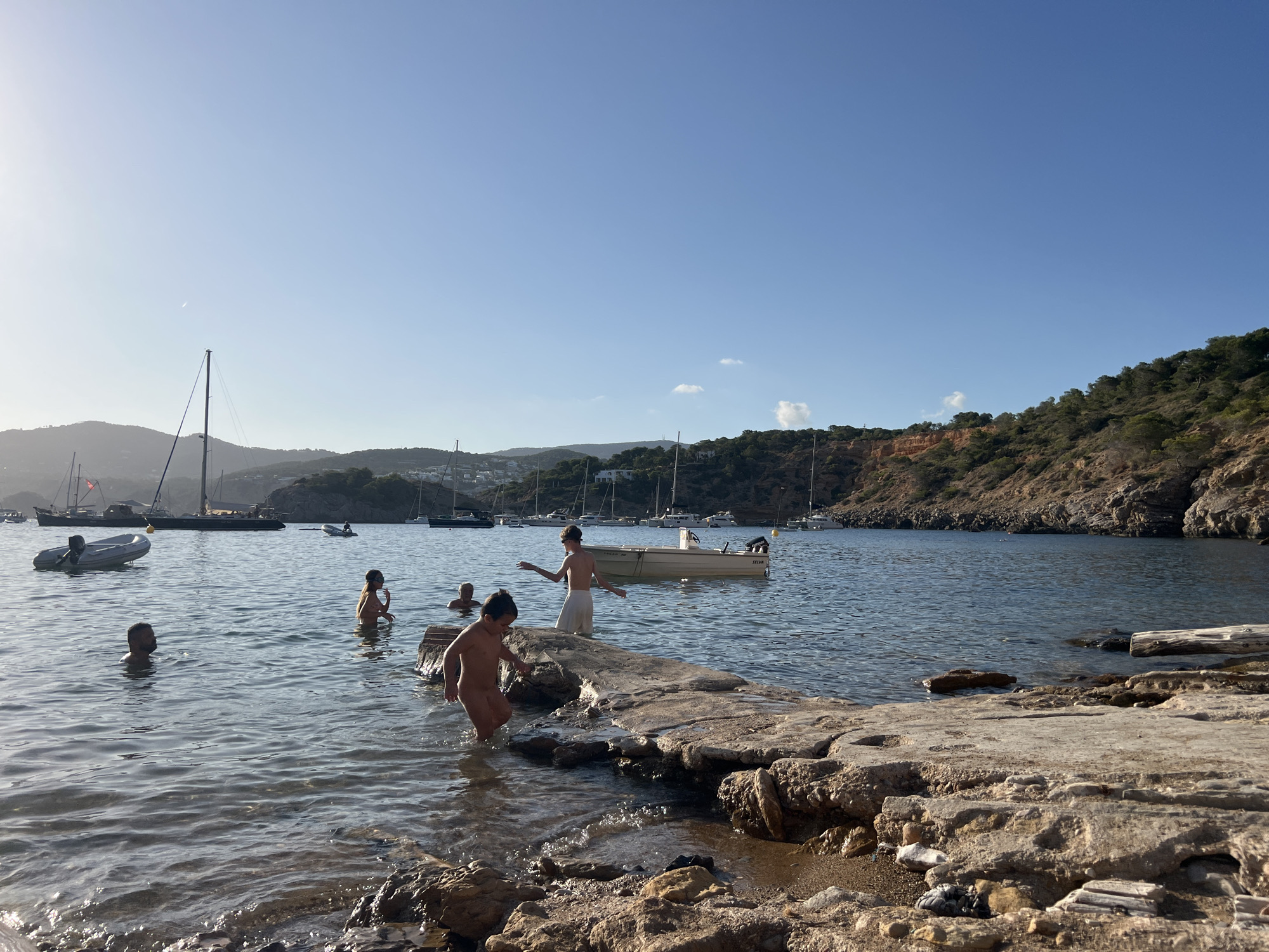The image depicts a sunlit bay with a rocky shoreline and calm, clear water. Several sailboats and smaller boats are anchored in the bay. A group of people are in the water, some swimming, and others standing on a low-lying rocky platform. One person, a child, is walking out of the water onto the rocks. Another child is standing on the rocks with a towel around their waist. In the background, a wooded hillside rises above the bay, dotted with houses. The sky is a clear, bright blue with a faint haze near the horizon. The scene suggests a warm, recreational day at the beach.