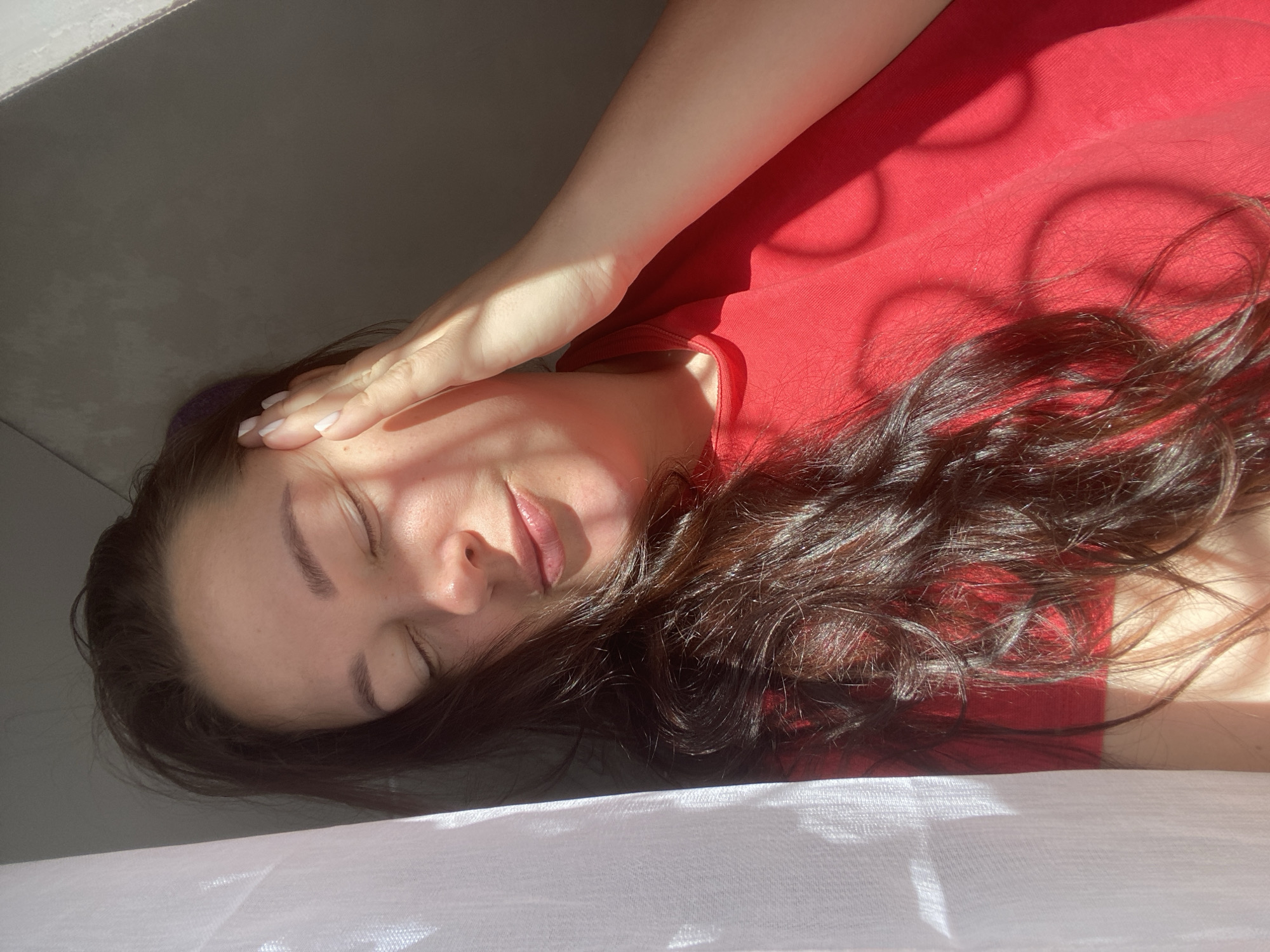 The image shows a woman lying down, likely indoors near a window in Sochi, Russia. She is wearing a red shirt and has dark, wavy hair. Her eyes are closed, and her hand is resting on her forehead, suggesting relaxation or perhaps mild discomfort. The room appears to be illuminated by bright sunlight, creating distinct shadows on her face and clothing. The background consists of a gray wall and what appears to be a white sheet or fabric in the foreground.