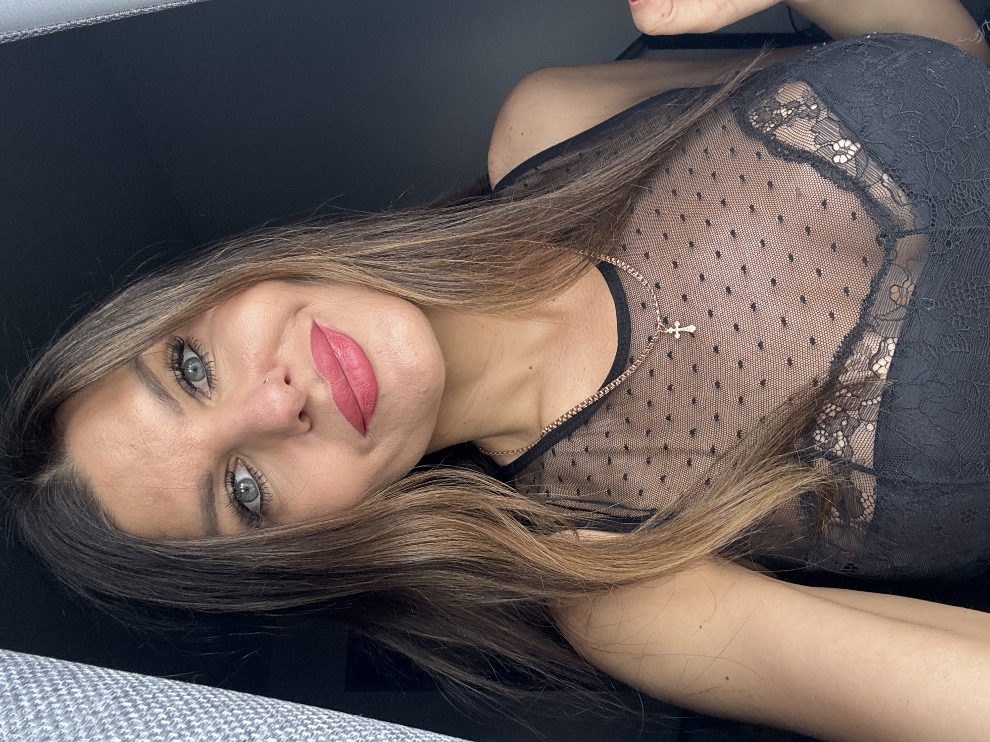 A woman with light brown hair and green eyes is lying down, looking at the camera. She is wearing a black sheer top with polka dots and lace details, and a gold necklace with a cross pendant. The background is a mix of dark gray and gray upholstery. The location is unknown, and the claim of it being in Chaikovsky, Russia cannot be verified from the image.