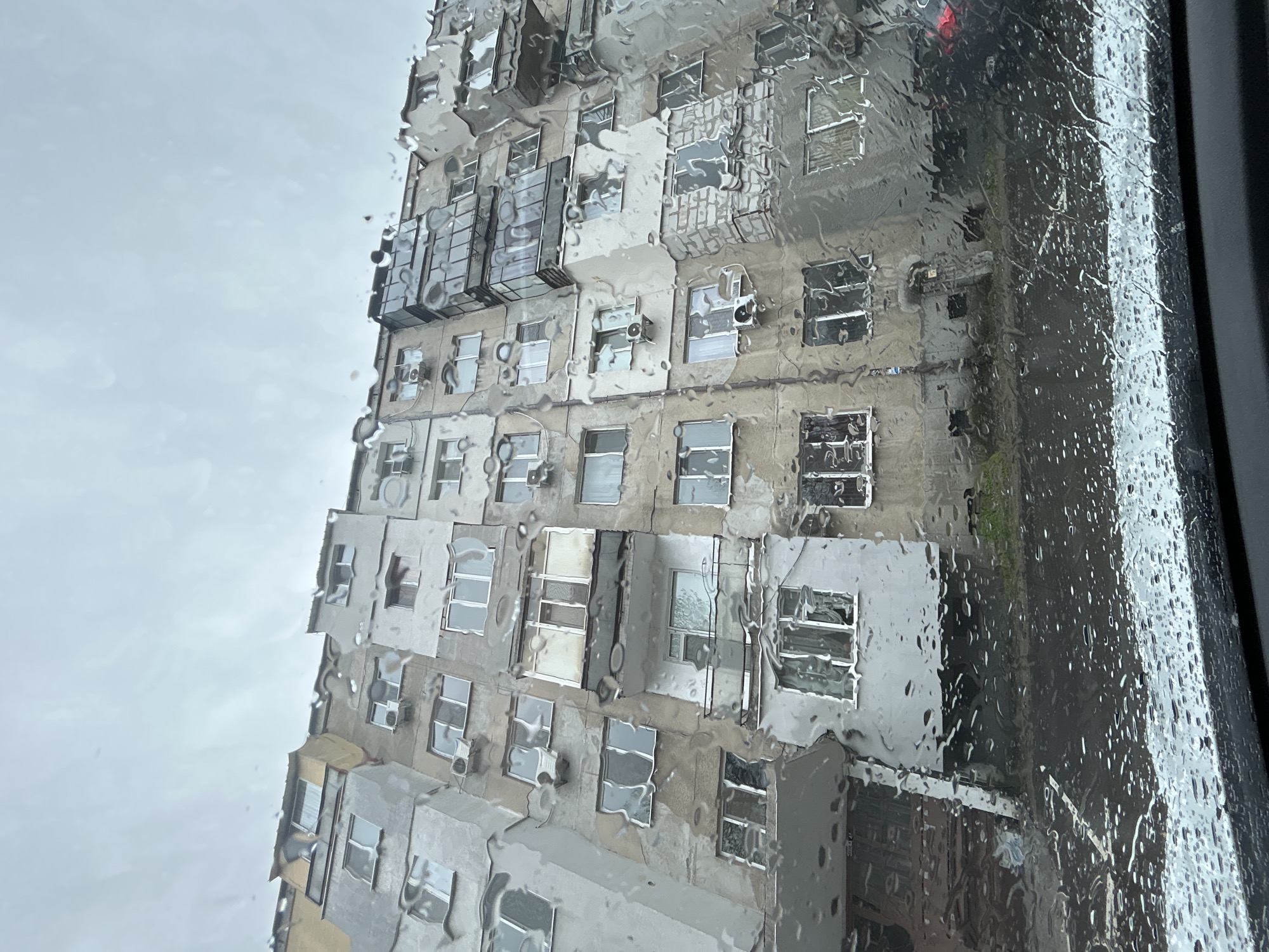 The image shows a tall, rectangular building with multiple stories, observed through a rain-streaked window. The building facade is characterized by numerous windows and protrusions, some of which appear to be air conditioning units or small balconies. The building's exterior has a weathered, aged appearance, with varying shades of beige and gray. The architecture seems utilitarian. The background sky is overcast and dull.
While the information to conclusively identify the building's location in Plovdiv, Bulgaria, is absent from the provided image and available data, based on the architectural style and visual clues the building could be in the eastern part of Plovdiv, Bulgaria, characterized by Soviet-era residential buildings, or in one of the city's older residential neighborhoods.
