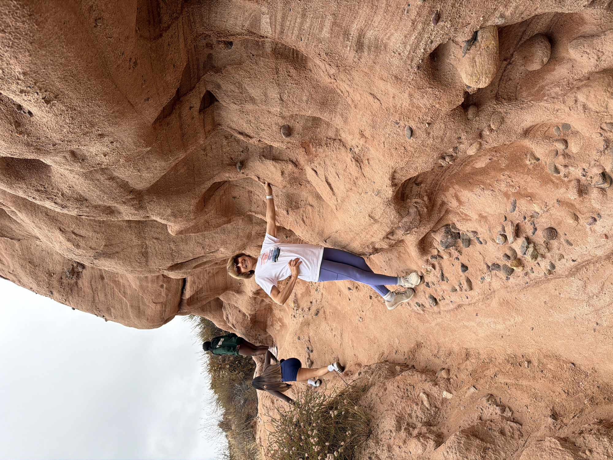 The image shows a woman in the foreground, standing on a dirt path next to a large rock formation. She is wearing a white t-shirt, blue leggings, and sneakers, and is smiling at the camera. In the background, two other people are walking up the path. The environment appears to be a desert or canyon area, with dry vegetation and red rock formations. The sky is overcast. Based on visual cues, the location is likely O'Neill Regional Park in Lake Forest, California, United States of America, known for its extensive network of hiking trails and sandstone canyons.