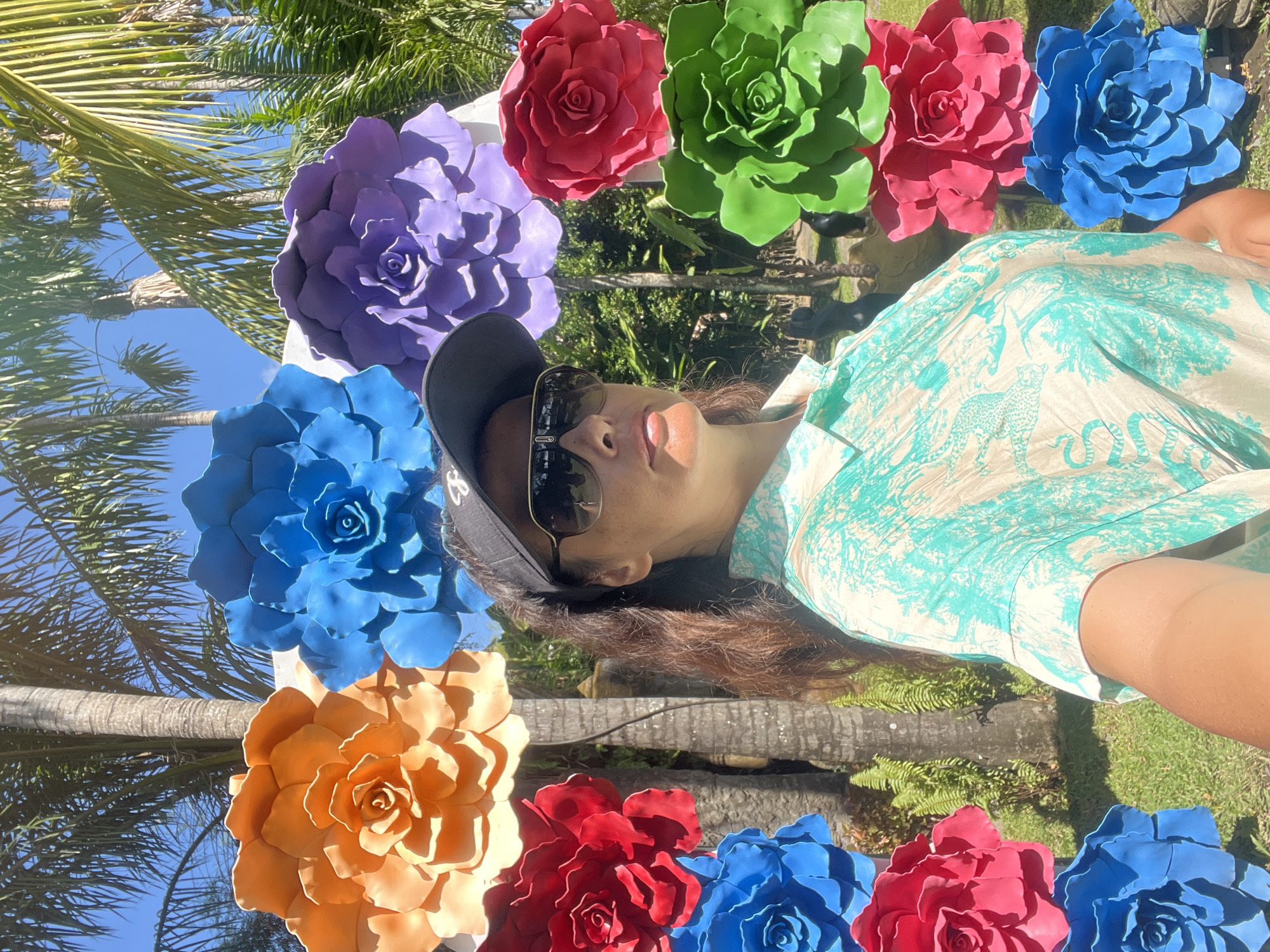 A medium shot captured from a low angle shows a woman taking a selfie in front of a decorative floral archway. The woman is wearing a black baseball cap, sunglasses, and a teal and cream patterned collared shirt. She is positioned in the foreground, her face angled towards the camera. The floral archway is composed of large, vibrant, artificial rose-shaped flowers in various colors, including orange, blue, purple, red, and green, arranged symmetrically. Behind the woman and archway, palm trees and lush green foliage are visible against a bright blue sky, suggesting an outdoor tropical setting. The overall scene conveys a cheerful and relaxed atmosphere. Based on visual cues such as abundant palm trees, lush vegetation, and the typical bright sunlight, the location is likely Denpasar, Indonesia, possibly in a park or garden setting.