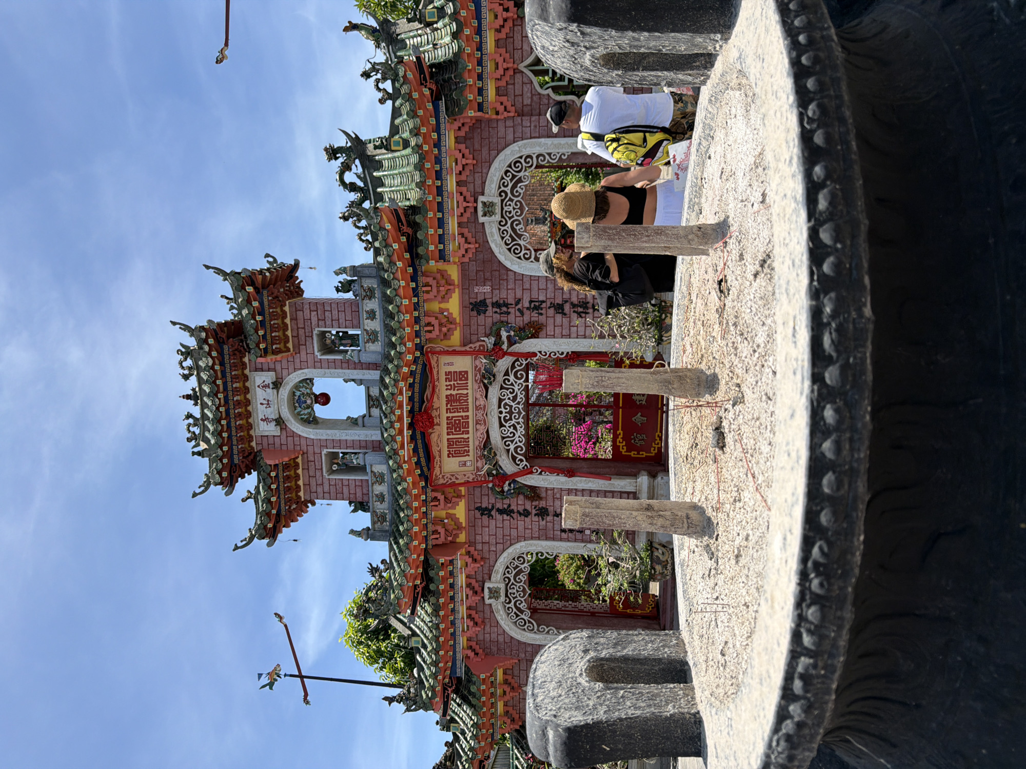 In Hội An, Vietnam, a wide shot captures the ornate facade of a temple, bathed in bright sunlight under a clear blue sky. The temple, painted in vibrant shades of pink and green, features intricate architectural details, including tiered roofs adorned with dragon motifs and decorative carvings. Red banners with Chinese characters hang across the entrance. In the foreground, a large, dark incense burner is filled with sand, with several incense sticks partially burned. Three people stand near the temple entrance, facing away from the camera. One person wears a black top and a straw hat, another a black t-shirt and a cap, and the third a white t-shirt and a hat. A green tree is visible behind the temple entrance. A weather vane shaped like a fish on a pole is positioned to the left of the temple. The overall scene depicts a cultural and historical landmark in Hội An.