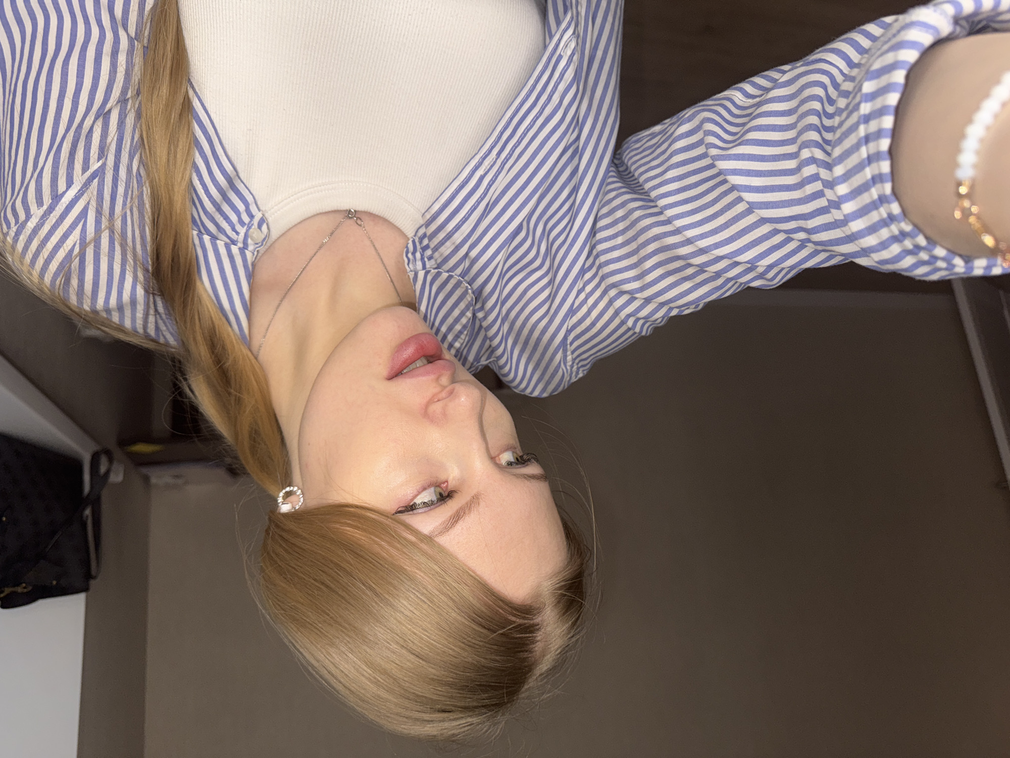 A blonde woman with fair skin is pictured indoors with a neutral background. She wears a white ribbed top and a blue-and-white striped button-down shirt. The woman has her hair in a ponytail tied with a decorative circular hair tie. A thin silver necklace is visible on her neck, and a white beaded bracelet is on her right wrist. A black handbag is visible in the background. There's no specific evidence to pinpoint the exact location as Perm, Russia.