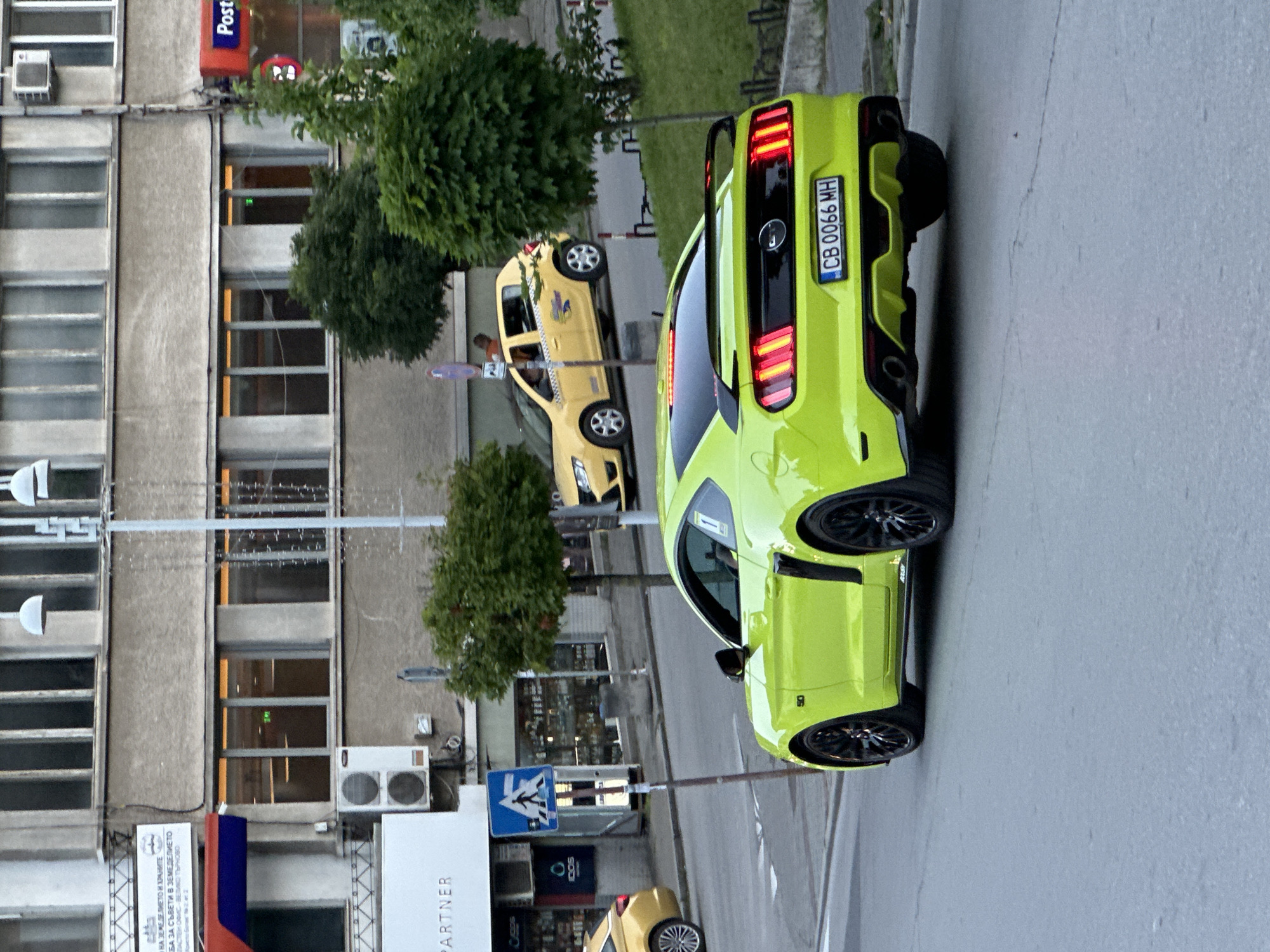 The image shows a lime green Ford Mustang parked on a city street in Veliko Tarnovo, Bulgaria. The car is in the foreground, viewed from the rear three-quarter angle. The car's license plate reads "CB 0066 MH". In the background, a yellow taxi is visible, along with a pedestrian crossing sign and buildings. The environment appears to be an urban setting with trees and streetlights. The weather is overcast. There are no people clearly visible interacting with the car or each other.