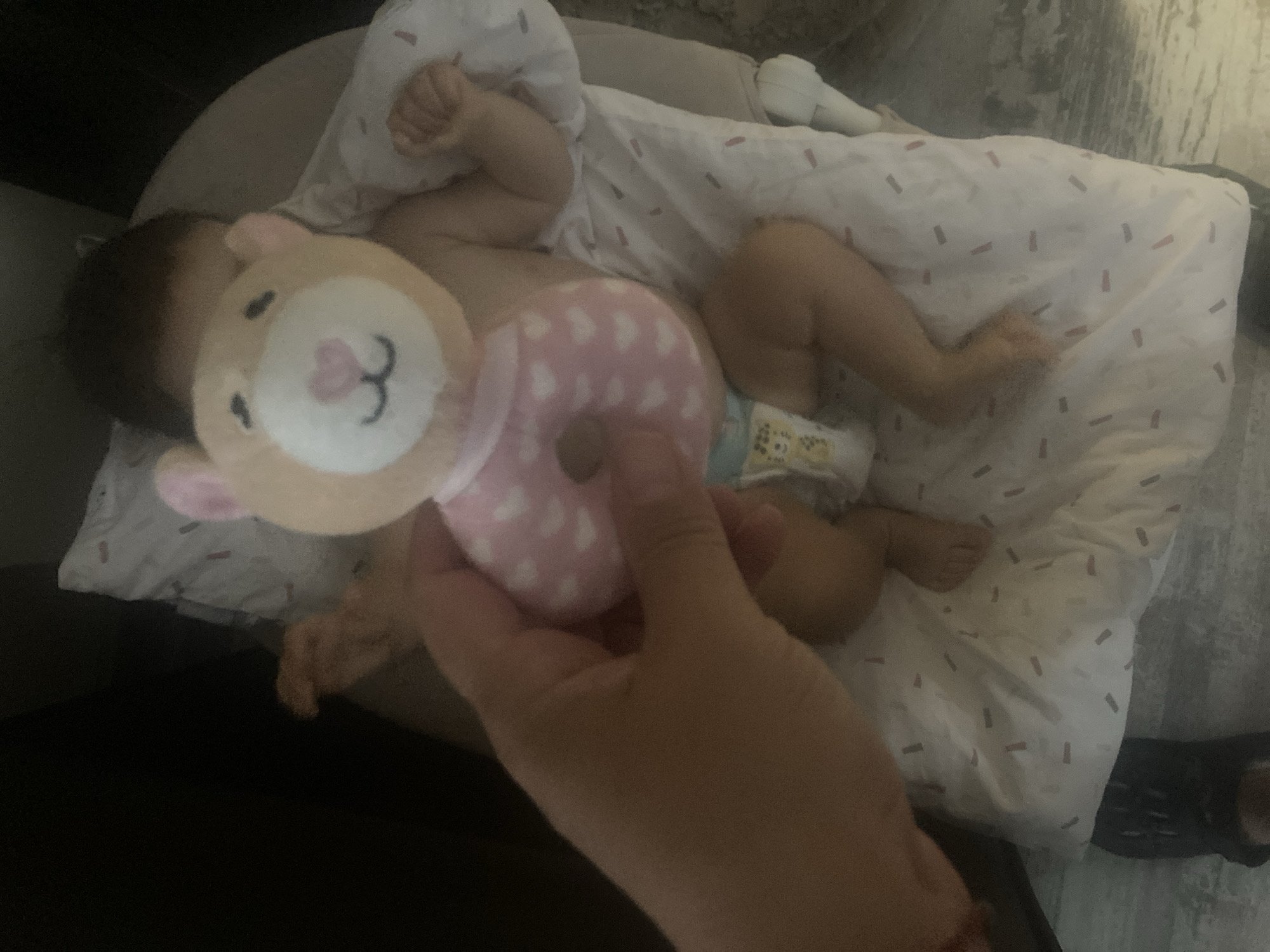 In an indoor setting, likely a nursery or bedroom, a person's hand holds a plush toy shaped like a bear's head with pink ears and a white face. The bear has a pink heart-shaped nose and black stitched eyes and mouth. The bear's body is a pink ring decorated with white hearts, and it appears to be a rattle or teether. The toy is positioned in front of a baby who is lying on their back on a white patterned blanket. The baby is wearing a diaper and is mostly unclothed. The blanket has a repeating pattern of small, thin, red and grey rectangular shapes. The background is dark, suggesting low lighting conditions. The scene does not provide any specific visual cues to determine a location within Burgas, Bulgaria.