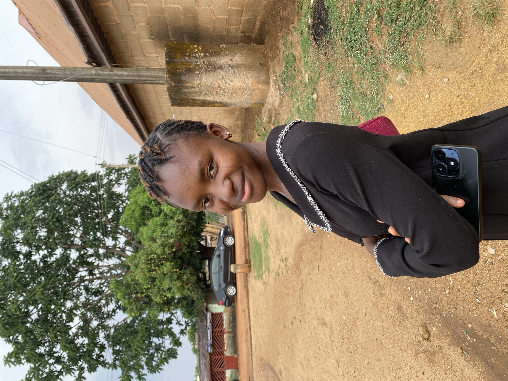 A young woman stands outdoors in Kuru Dungus, Nigeria. She is holding a smartphone and smiling at the camera. The location appears to be a residential area with a dirt road. A building, a tree, and a car are visible in the background. The woman is wearing a black outfit. A red purse is visible as well.