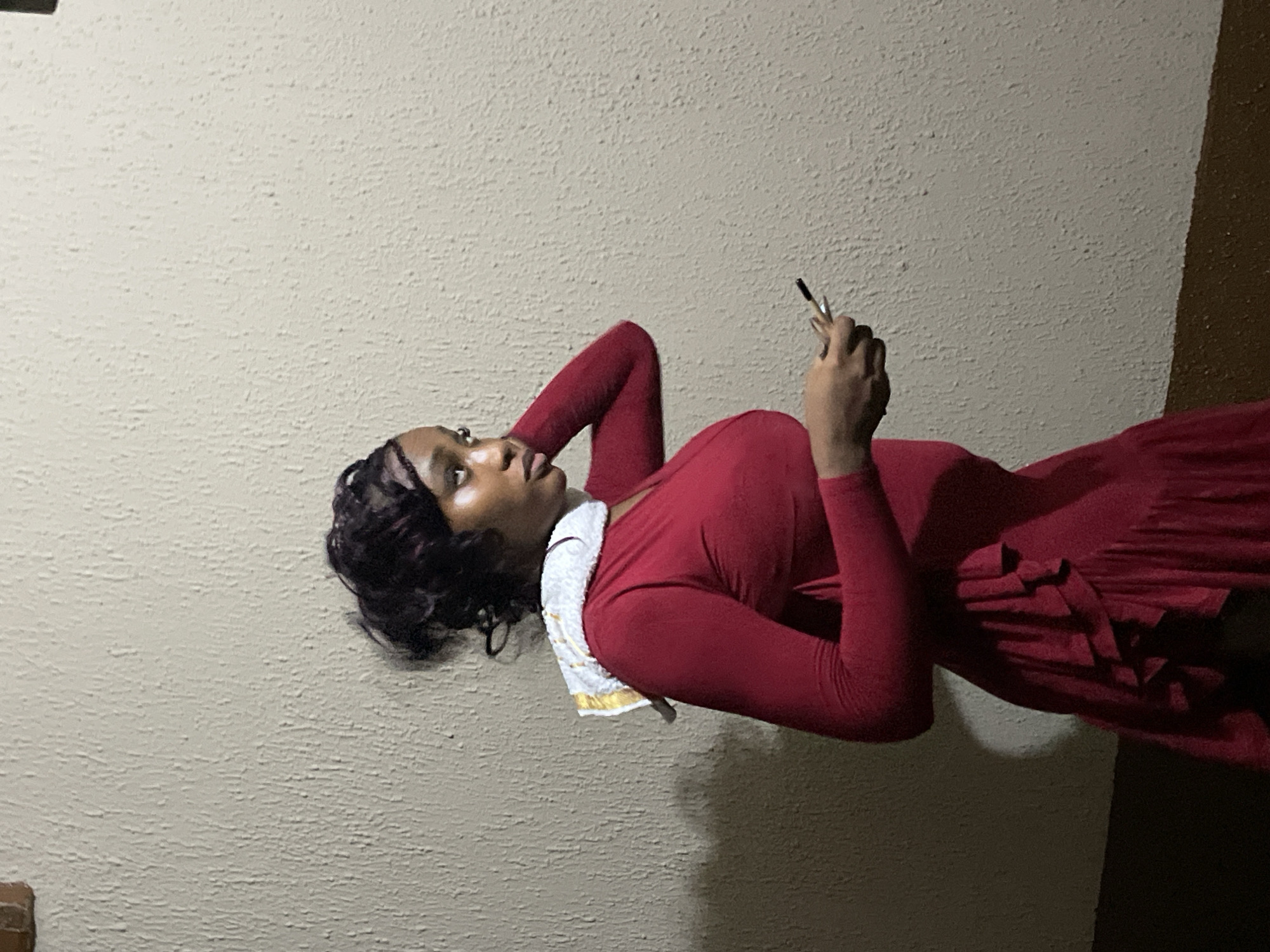 A young Black woman in a red dress reclines against a textured, light-colored wall. She holds a thin object, possibly a pen or cigarette, in her right hand, and looks towards the upper left corner of the frame. A white cloth with gold trim is draped around her neck. The background is a wall, and a darker area is visible at the edge of the frame, suggesting a doorway or corner of a room. There is no evidence to confirm that the image was taken in Ijegun, Nigeria.