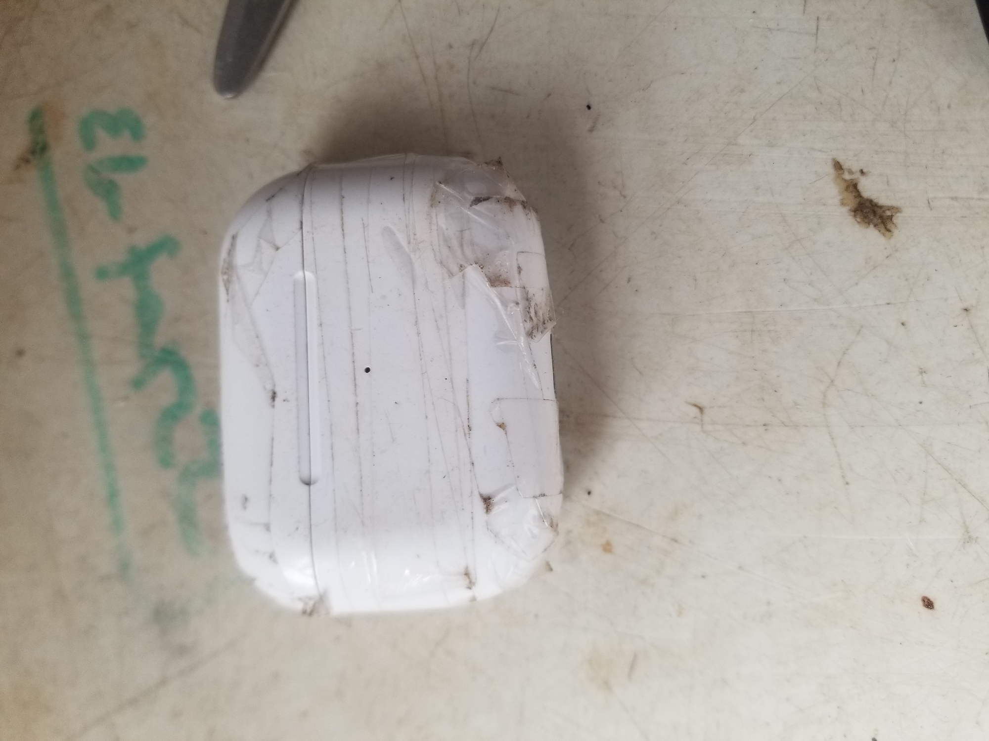 In Jauro Yinu, Nigeria, a white charging case for wireless earbuds, possibly AirPods, rests on a light-colored, textured surface. The case is heavily wrapped in clear tape, which appears to be applied haphazardly and is creased and soiled. Some of the tape has pulled away, revealing a small amount of dirt and debris on the case itself. The surface beneath the case is marked with numerous scratches and faint green markings that appear to be graffiti or a similar inscription. The overall impression is one of an object that has been crudely repaired or sealed for storage or transport.