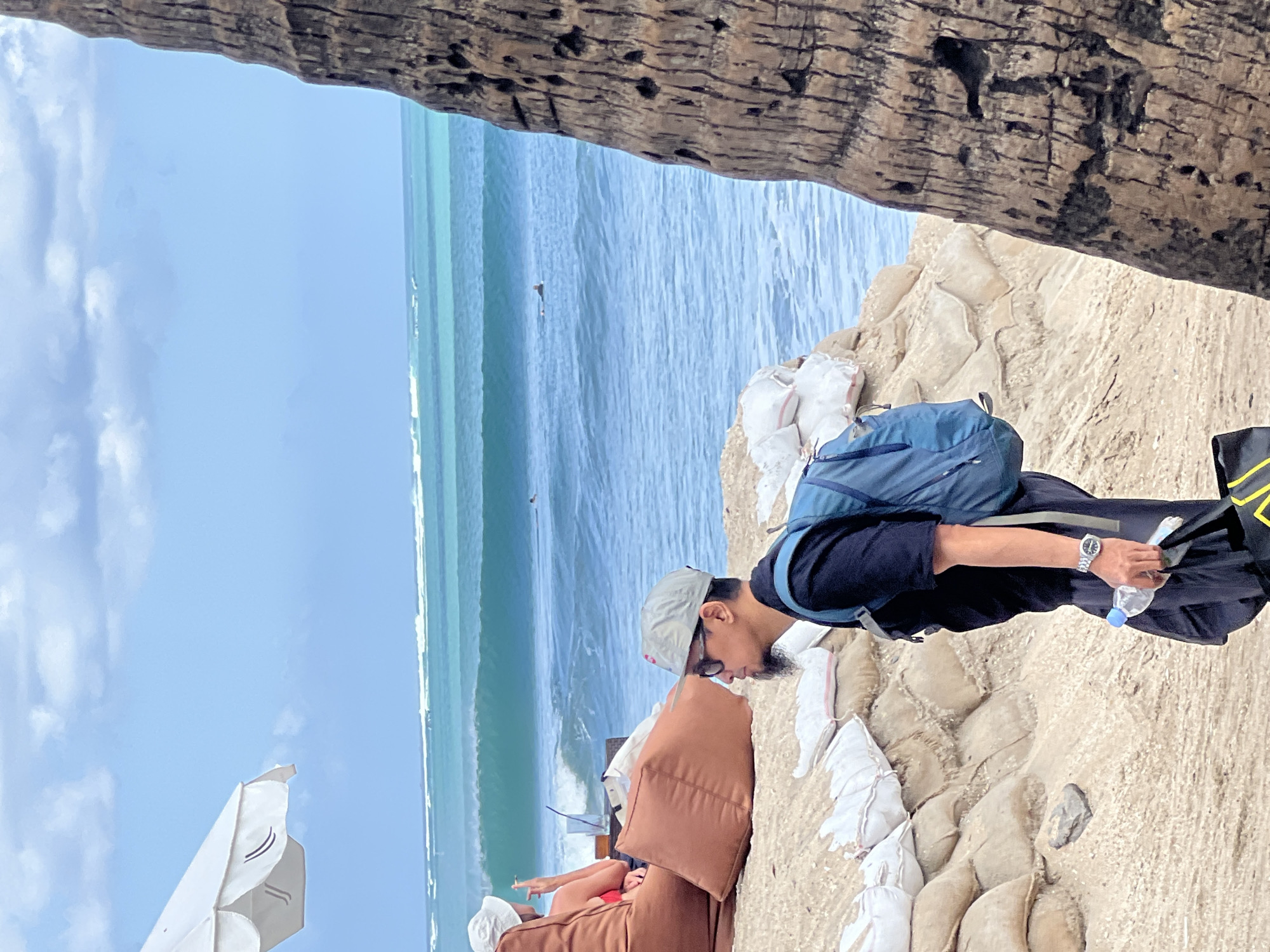 A man wearing a black t-shirt, dark pants, a grey baseball cap, and sunglasses, with a goatee, walks along a sandy beach in Kuta, Indonesia. He carries a blue backpack, a plastic bottle of water in his right hand, and a black bag with yellow straps in his left. In the background, waves crash on the shore, and a few surfers are visible in the turquoise water. A person in a white hat sits on beanbag chairs under a white umbrella to the left, smoking a cigarette. Sandbags are stacked along the beach, possibly to prevent erosion. A palm tree trunk is visible on the right side of the frame. The sky is partly cloudy.