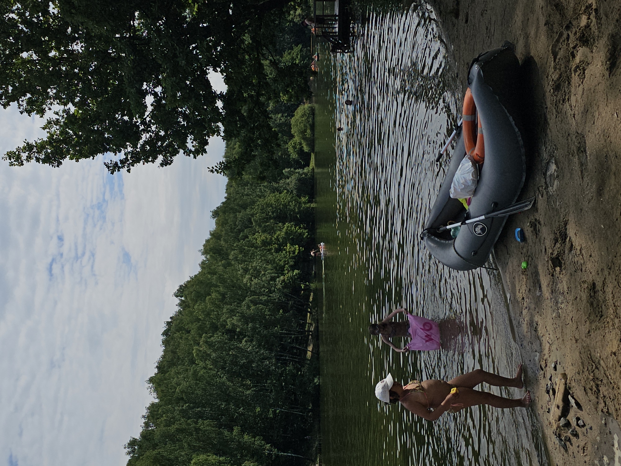 In this vertically oriented photograph, a group of people are enjoying a day at a sandy beach and lake. The scene is set on a warm, sunny day, likely during the summer, with a bright blue sky dotted with white clouds visible above the lush green trees that line the opposite bank of the lake. The water is calm and appears inviting, with ripples reflecting the sunlight.
In the foreground, a woman wearing a bikini and a white cap stands partially in the water, holding a yellow object. To her left, another person, partially submerged in the water, is holding a pink towel or cloth. Further back in the water, other individuals can be seen swimming and relaxing.
On the sandy shore to the right, a deflated inflatable boat lies on its side. An orange life preserver is placed on top of the boat, and two paddles are visible. A plastic bag and a small blue object are also near the boat.
In the background, across the lake, is a dense forest of green trees. A wooden dock or pier extends into the water, with several people visible on it and near it. Some are in small boats, possibly rowing or enjoying the water. The overall atmosphere of the image is one of leisure and recreation. The image appears to be taken in Moscow, Russia. There is no visible text in the image.