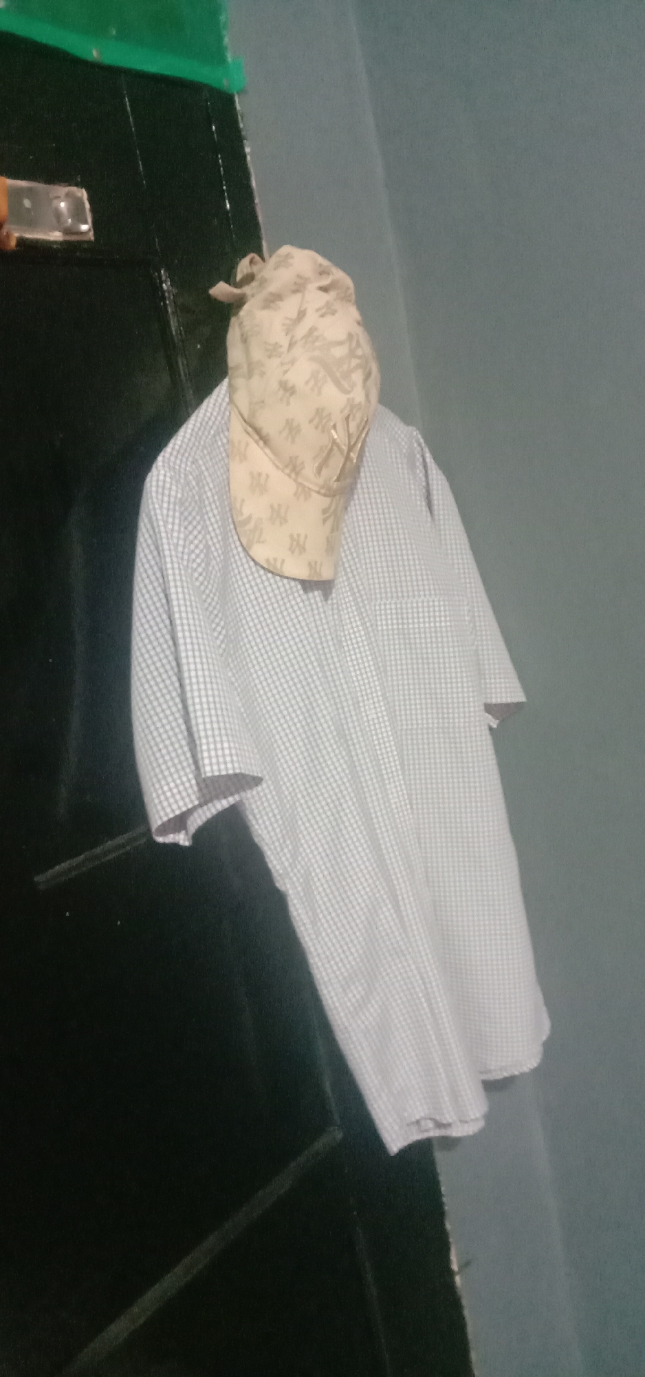 A beige baseball cap with a repeating "NY" logo pattern is placed on top of a light blue and white checkered short-sleeved collared shirt. The shirt and cap are hanging on what appears to be a dark door or closet. The background consists of a teal-colored wall. The lighting suggests an indoor setting, likely a home, with no specific identifiable landmarks pointing to Kendari, Indonesia.