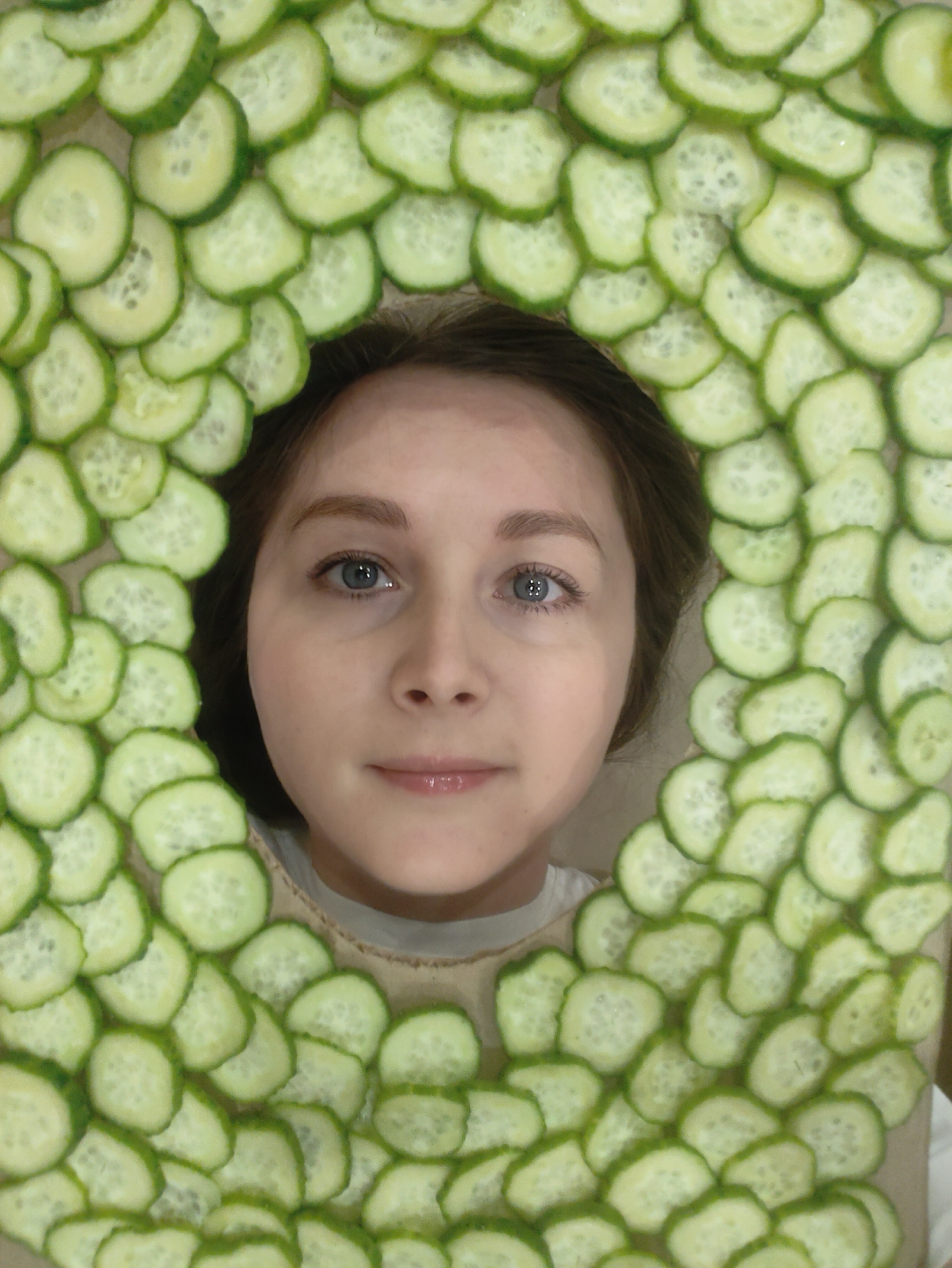 A person with fair skin, blue eyes, and brown hair is centrally framed within a rectangular border composed of overlapping sliced cucumbers. The cucumber slices are green with lighter-colored interiors containing seeds, and they create a frame that surrounds the person's face, from the forehead down to the collarbone area. The person is wearing a light-colored shirt. The location is unknown.