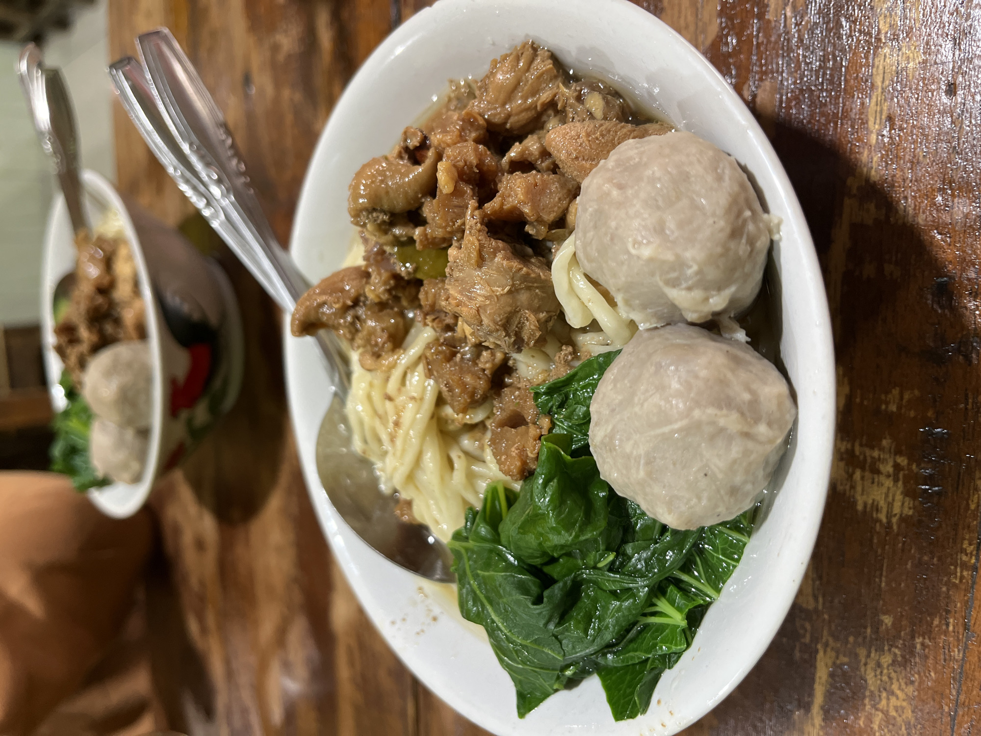 A close-up, eye-level shot shows two bowls of Indonesian chicken noodles with meatballs on a dark, wooden table. The bowl in the foreground, which is white with a colorful rim, contains a generous portion of noodles topped with savory chicken pieces, a side of dark green leafy vegetables, and two large, light brown meatballs. A silver spoon rests in the noodles. A second bowl, similar in design, is visible in the blurred background, also containing noodles, chicken, and meatballs. The lighting is warm and natural, highlighting the textures and colors of the food. The scene suggests a casual dining experience, likely in a restaurant in Sidoarjo, Indonesia, known for its culinary offerings.