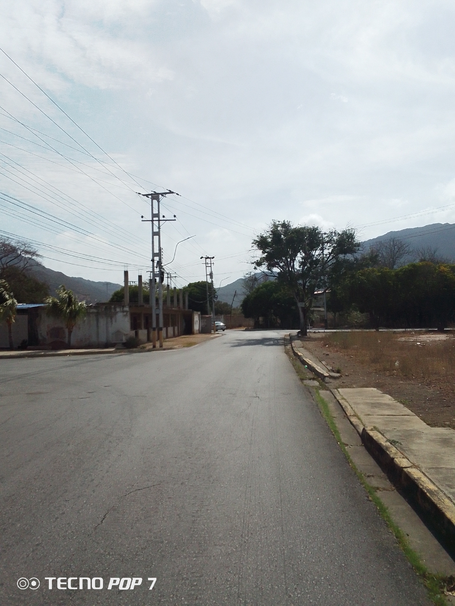 The image shows a street in Pedro González, Venezuela. The road is paved and has a sidewalk on the right side. There is a partially constructed building on the left side of the street. Power lines run overhead, supported by utility poles. Vegetation is visible on both sides of the road, including trees and dry grass. In the distance, there are hills or mountains under a cloudy sky. A car is parked on the side of the road further down the street. The image was taken with a TECNO POP 7 phone.