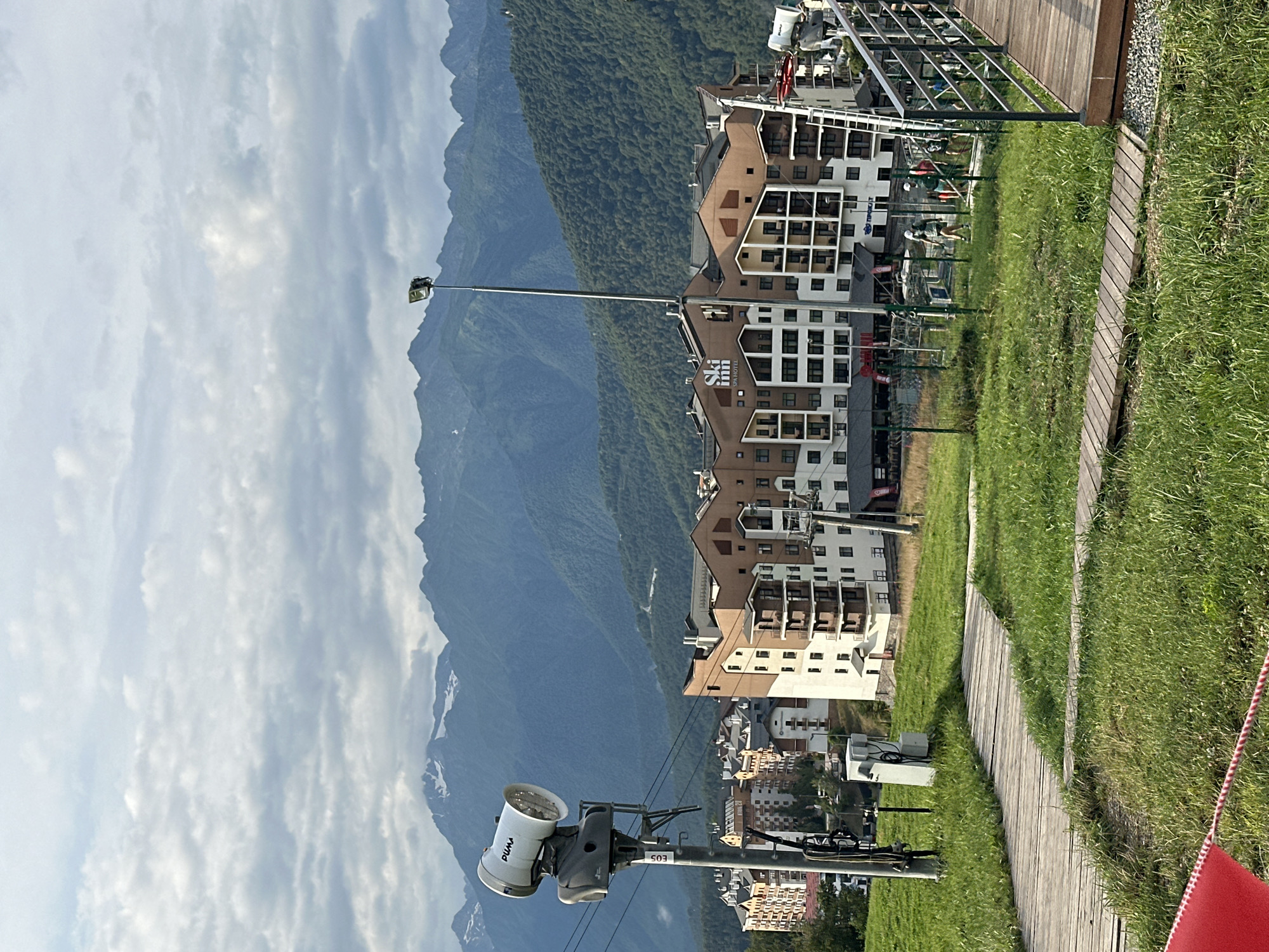 The image captures a scenic view of a mountain resort in Estosadok, Russia. In the foreground, a wooden walkway meanders through a grassy area. To the left, a snow cannon is mounted on a pole, equipped with a fan and a nozzle, indicating its potential use for snowmaking. In the middle ground, several multi-story buildings with distinctive brown and white facades are visible, likely hotels or residential complexes, with balconies and numerous windows. A street light on a tall pole stands prominently between the buildings and the mountains. The background is dominated by a majestic mountain range with lush green slopes leading up to snow-capped peaks under a cloudy sky. Several people are scattered on a path near the buildings, some appearing to be walking or participating in an activity. The overall scene suggests a resort environment, possibly during the off-season or in preparation for winter activities.