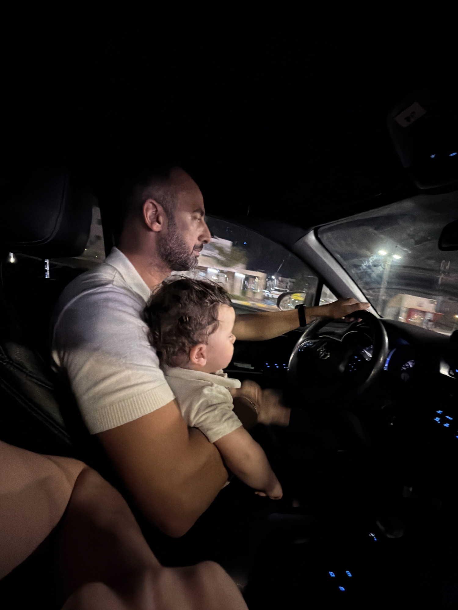 An adult male with a beard and a white polo shirt is driving a car at night. He is holding a young child on his lap, who is wearing a white shirt. The child is looking to the right. The driver's hands are on the steering wheel. The interior of the car is dark, with some illuminated buttons on the dashboard. Outside the car, there are blurred lights that suggest the vehicle is moving through an urban environment. The scene appears to be in Sharm El Sheikh, Egypt, given the presence of what looks like a Toyota logo on the steering wheel, a brand commonly found in Egypt.