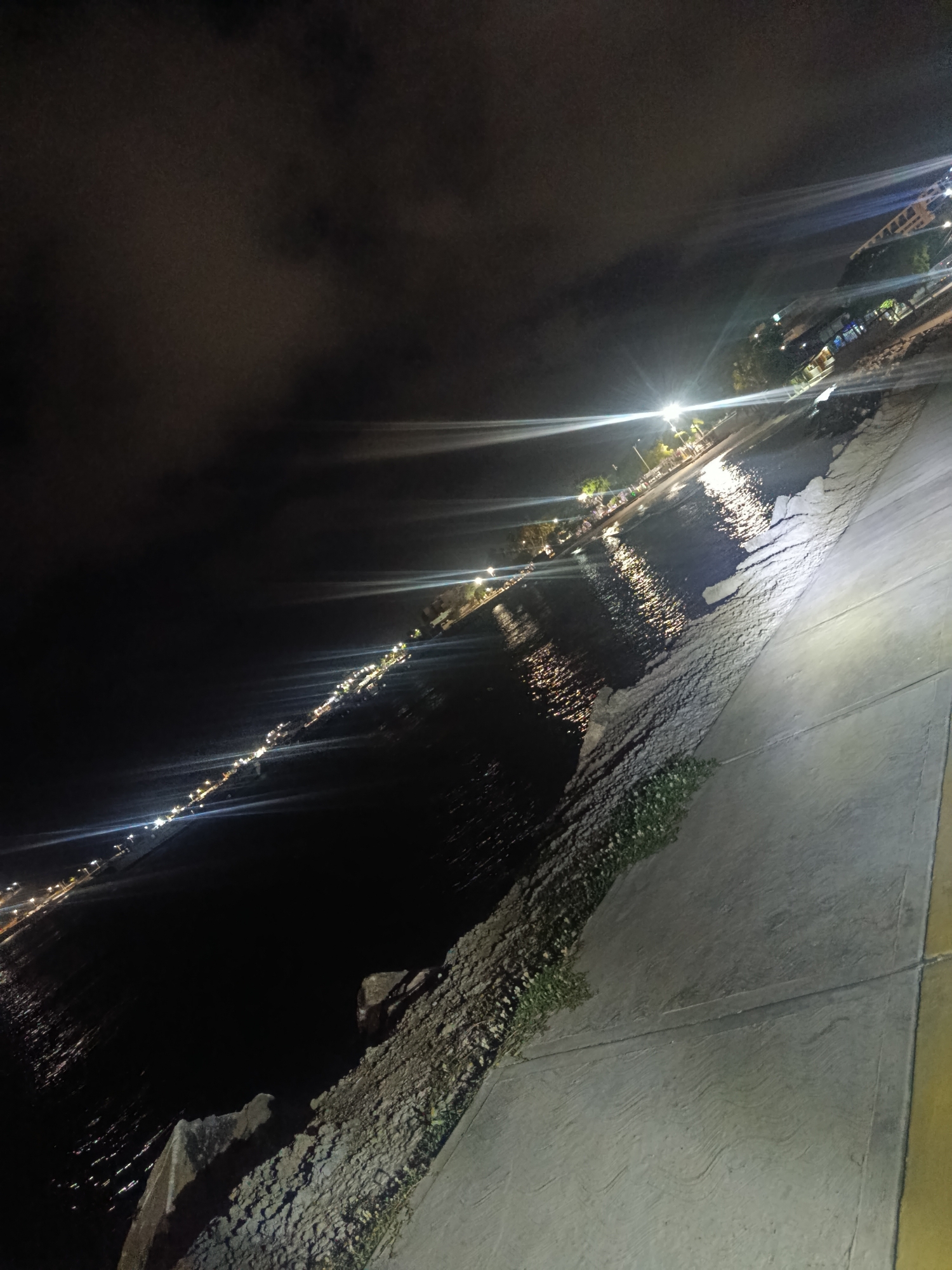 The image depicts a nighttime scene along a waterfront in Juan Griego, Venezuela. The perspective is from a paved walkway, angled downwards towards the water. The walkway is textured with wavy patterns. To the left of the walkway, there is a rocky embankment with some sparse vegetation growing on it. Beyond the embankment lies the dark water of the sea, reflecting the lights of a distant town or promenade. The town is lined with numerous small, bright lights, suggesting buildings or streetlights. These lights create streaks and reflections on the water's surface due to the long exposure of the photograph. The sky is dark and cloudy, with some light sources causing lens flares and visible light rays radiating outwards, indicating bright artificial lights in the distance. There are no people or specific actions visible in the image, just the static elements of the urban waterfront at night.