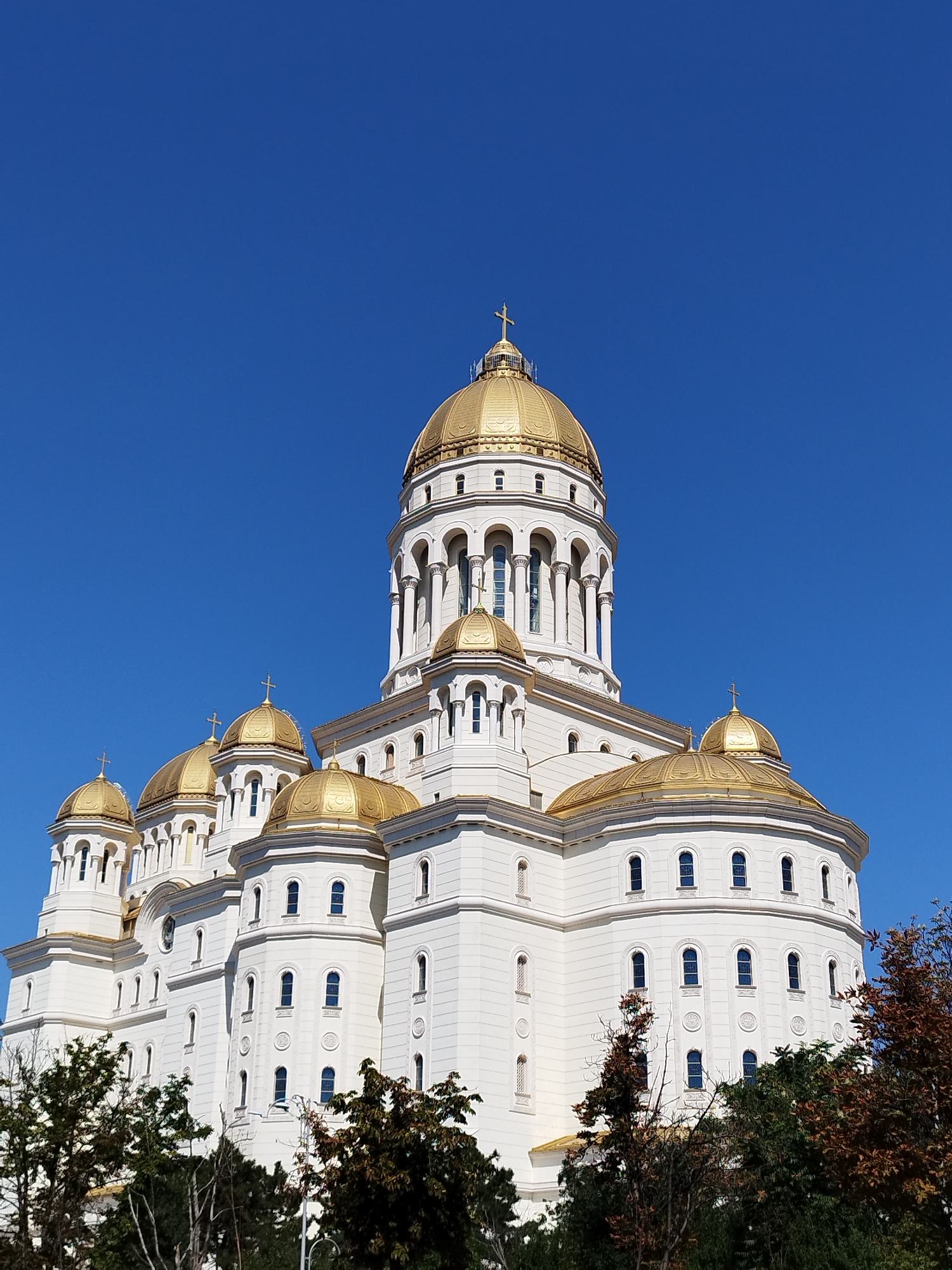 The image shows the People's Salvation Cathedral, a Romanian Orthodox cathedral located in Bucharest, Romania. The cathedral is a large, white building with multiple golden domes, the largest of which is at the center and topped with a cross. Smaller golden domes surround the main dome, and the building features numerous arched windows. The sky is a clear, deep blue. In the foreground, the tops of several trees with green and brown foliage are visible.