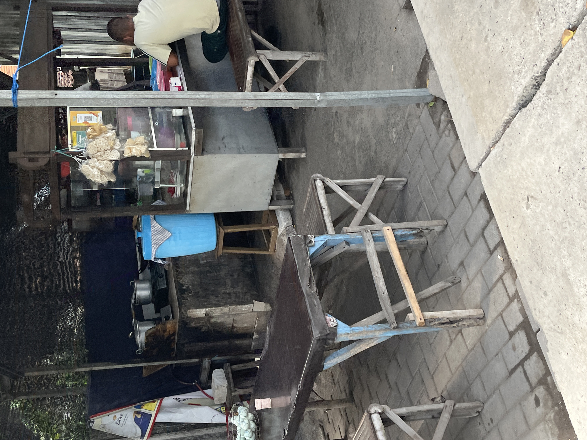 This image shows a street food stall located in Kepatihan, Indonesia. In the foreground, a dark brown rectangular table with a blue wooden frame and a wooden bench are set up on a paved walkway. In the background, a food stall is visible with a person seated on a wooden bench, facing away from the camera. The stall has a glass display case filled with food items, including bags of crackers hanging from the ceiling. Cooking equipment, such as pots and a gas stove, is also present. A large blue plastic container sits next to the stall. The environment appears to be an outdoor setting, with a roof providing shade over the stall.