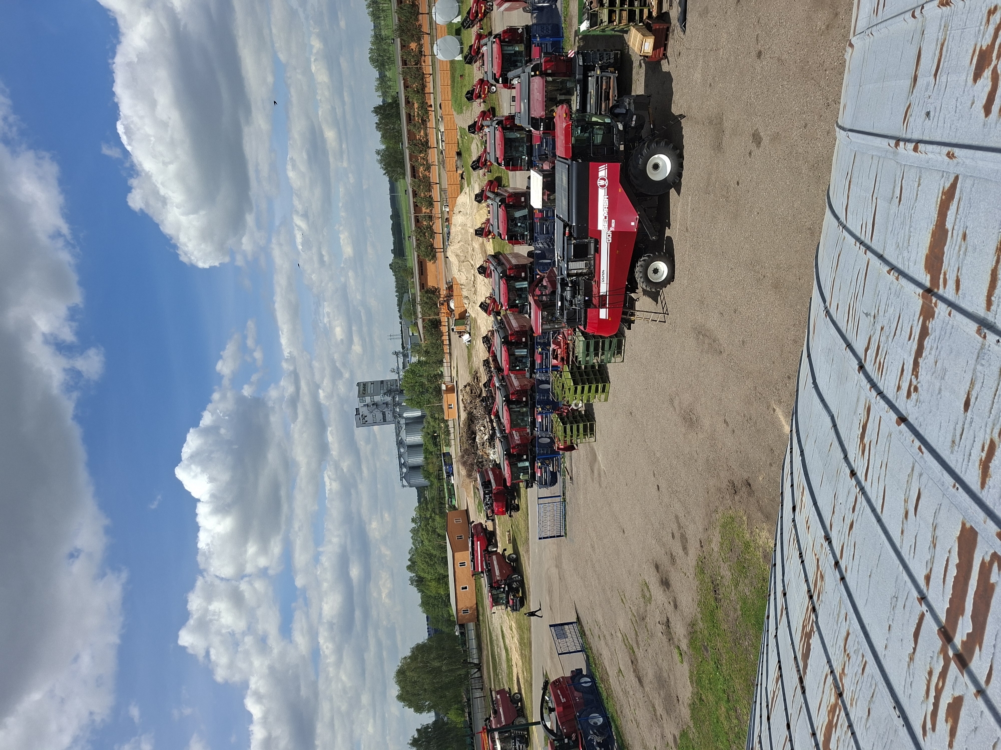 The image depicts an open-air storage or sales yard in Zhabinka, Belarus. The yard is filled with a large number of agricultural vehicles, predominantly red combine harvesters and tractors. Several appear to be new, possibly awaiting sale or distribution. The vehicles are densely packed on a gravel or concrete surface.
In the background, there are metal grain silos, buildings, and some greenery. The sky is visible, displaying partly cloudy conditions with a blue sky. A section of the top of a corrugated metal silo is visible in the foreground.
There is a single visible person walking along the edge of the yard.