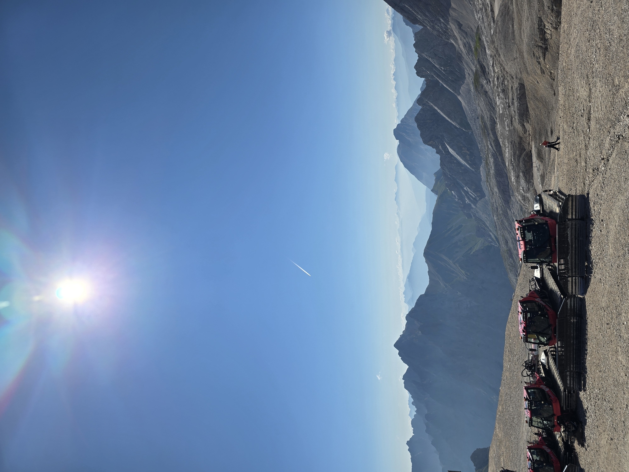 The image depicts a panoramic view from a high altitude, likely the Zugspitze, near Garmisch-Partenkirchen, Germany. The sky is a clear, vibrant blue, with the sun shining brightly and causing a lens flare. A contrail from an airplane is visible high in the sky. In the foreground, a line of red snowcats, also known as PistenBully vehicles, are parked on a gravelly surface. These vehicles are typically used for grooming ski slopes. Further back, a vast mountain range stretches across the horizon, with layers of peaks fading into a hazy blue in the distance. The terrain in the mid-ground appears rocky and barren, suggesting a high-alpine environment. One person, dressed in red and black, is seen walking away from the snowcats, possibly carrying a broom, while another person is visible further in the distance on the left. The overall scene is one of immense natural beauty and the presence of specialized equipment for mountain activities.