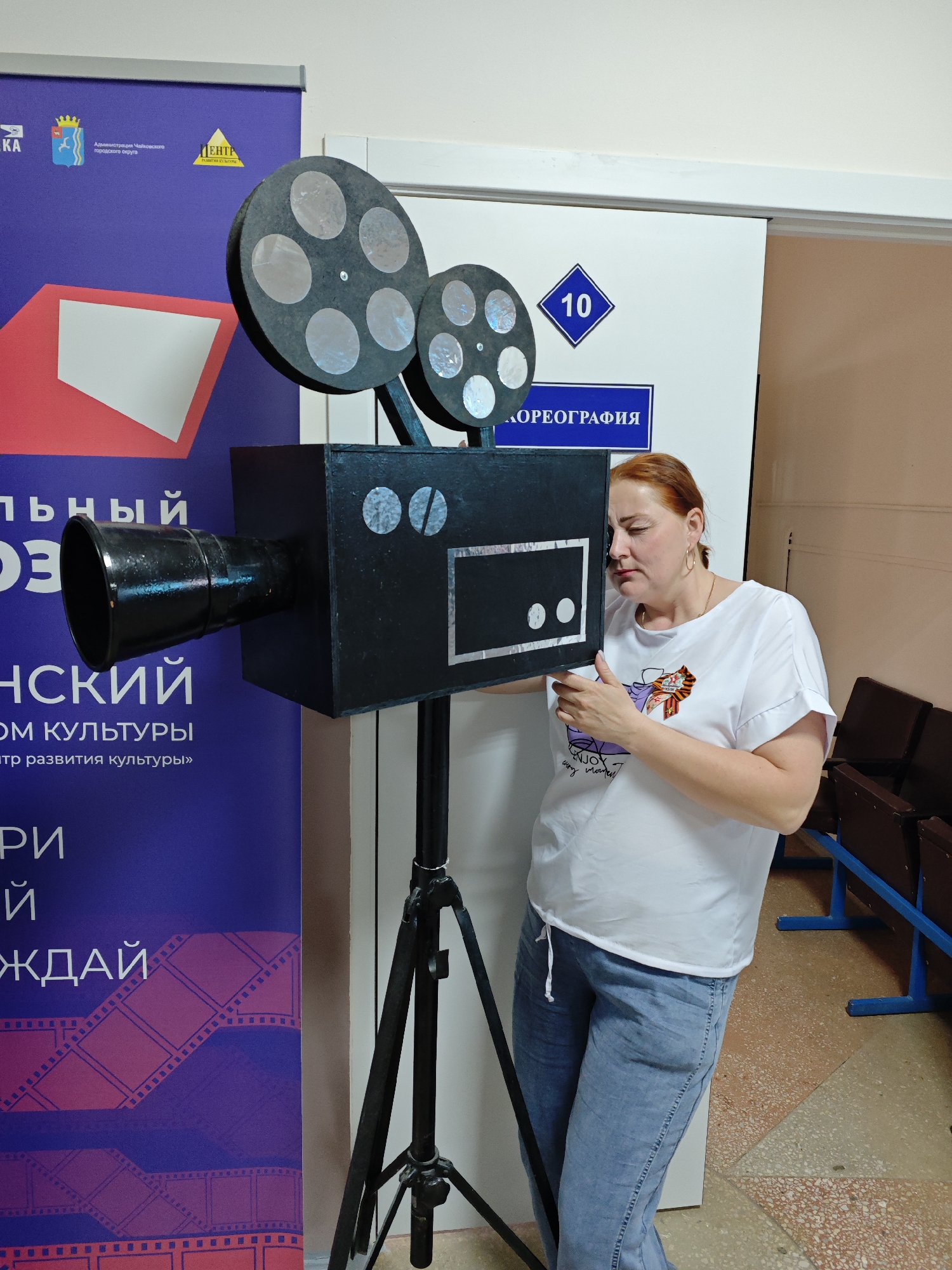A woman is standing indoors next to a black movie camera prop. The camera is on a black tripod. To the left of the camera is a purple banner with text. A door with a blue sign is behind the camera. The woman is wearing a white t-shirt and blue jeans. Benches with blue frames are behind her. The text on the banner says "Администрация Чайковской городского округа," "Центр," "10," "ХОРЕОГРАФИЯ," "льный," "ОЗ," "НСКИЙ," "ОМ КУЛЬТУРЫ," "тр развития культуры," "РИ," and "ЖДАЙ." The building is located in Фоки, Russia.