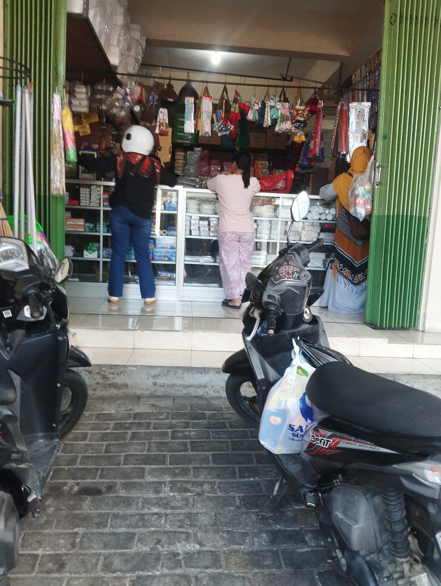 A person wearing a white helmet and a black jacket is standing in front of a store in Malang, Indonesia. The person is facing away from the camera and is surrounded by various items on display. The store appears to be a retail establishment selling a variety of goods, including what look like household items, clothing, and possibly food products. The store has green accordion-style doors on the right side, which are partially open. In the foreground, two motorcycles are parked, one on the left and one on the right, partially obscuring the view of the store entrance. The ground is paved with grey bricks. Another person, wearing a light pink shirt and patterned pants, is standing inside the store, facing towards the shelves of merchandise. A third person, wearing a yellow headscarf and a patterned dress, is also inside the store, near the entrance.