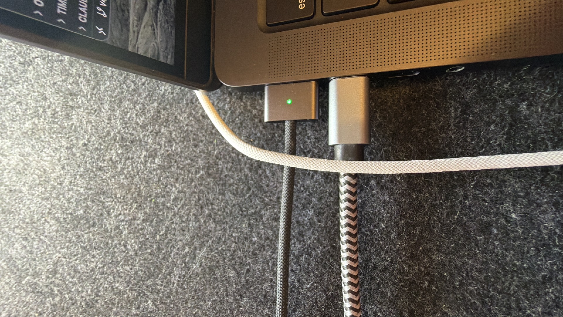The image shows a close-up of a laptop with two cables plugged into a port. The laptop's keyboard is partially visible at the top of the frame. One cable is a USB-C cable, connected to a silver adapter with a green LED light indicating it is powered on. The second cable is also a USB-C cable, with a black and white chevron pattern, plugged in next to the adapter. Both cables are connected to the side of the laptop, which rests on a textured, dark grey surface, likely a desk or table covered with felt material. The scene is indoors, with natural lighting suggested by the illumination of the adapter's LED. No people are visible in the image. The overall impression is one of a functional setup, with devices being connected or charged.