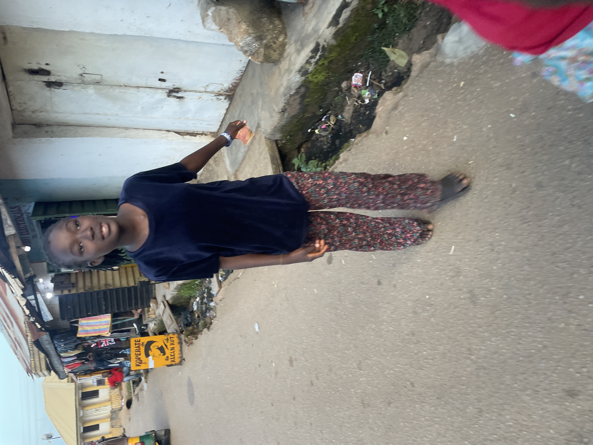 In an outdoor scene set in Jos, Nigeria, a young Black female, appearing to be in her early to mid-teens, walks on a paved road. She is wearing a dark blue t-shirt and patterned pants. Her left hand, adorned with a blue and white beaded bracelet, is extended, holding what appears to be a small object, possibly currency. Her bare feet are visible, and she is looking towards the camera with a neutral expression.
The background shows a street lined with shops. One shop has clothing for sale displayed outside, and a yellow sign with a silhouette of a man's head and the text "KOPERATE" and "KLEAN KUT" is visible. Another building to the right has a large, weathered white door with visible locks. A dirty, open drainage channel runs alongside the road, containing visible debris. The overall environment suggests a bustling, possibly informal commercial area in Jos. A part of a yellow and green auto-rickshaw is visible on the far left of the image.