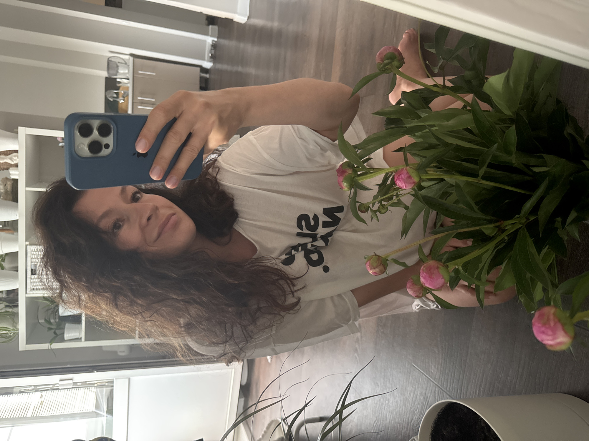 The image shows a young woman with long, dark, curly hair taking a mirror selfie in an indoor environment. She is wearing a white t-shirt with a black graphic design and is seated on a dark wooden floor. In the foreground, there is a large potted plant with several pink peony buds and green leaves. A white pot is visible to the left of the frame. In the background, a white shelving unit filled with plants and decorative items is against a wall. The room appears to be a living space or kitchen, with a countertop and cabinets visible in the distance. The overall scene suggests a casual, domestic setting. The image does not provide any specific details to determine the location in the city of Chaikovsky, Russia.