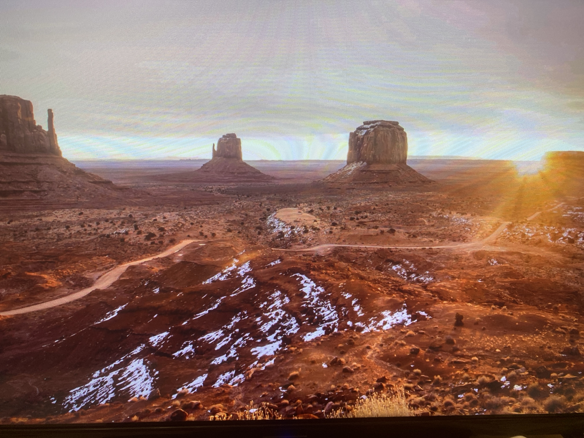 This media file is a wide shot of a vast desert landscape, likely Monument Valley, captured from a slightly elevated vantage point. The scene is dominated by several iconic rock formations (buttes and mesas). To the far left, a tall, slender rock pinnacle stands prominently, part of a larger formation. In the mid-ground, two distinct, large buttes resembling mittens are visible: one to the left and another to the right, both featuring flat tops and steep, eroded sides. A bright, warm light source, characteristic of either sunrise or sunset, emanates from the right side of the horizon, casting a golden glow and long shadows across the terrain.
The ground is a rich reddish-brown, typical of desert soil, and is uneven with sparse vegetation, appearing as small shrubs and clumps of grass. Notably, there are numerous patches of white snow scattered across the landscape, especially in the foreground and on the slopes of the formations, indicating cold weather, possibly winter or early spring. Several unpaved dirt roads or tracks wind through the desert floor, guiding the eye into the distance. The sky is hazy with soft, diffused light, gradually transitioning from a brighter, lighter tone near the horizon to a slightly darker shade overhead. The overall atmosphere is majestic and serene. No people, objects other than natural formations, or visible text are present. The image quality suggests it is a photograph taken of a screen, evidenced by the visible pixel grid and a dark border at the bottom.