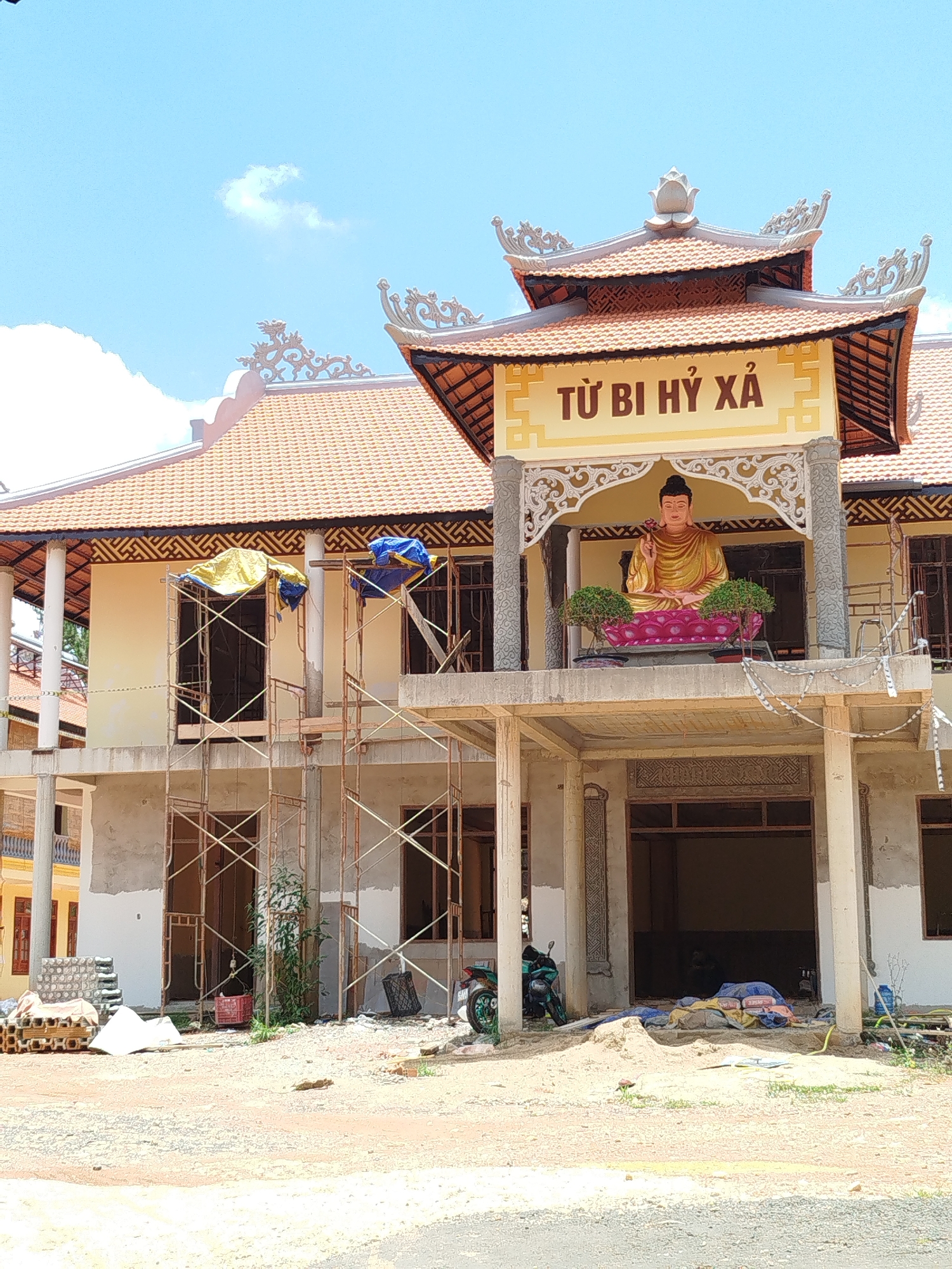This image captures a multi-story building, likely a Buddhist temple or pagoda, in the midst of construction or renovation under a bright, clear blue sky with sparse white clouds. The upper level of the building showcases traditional Vietnamese architecture, featuring an ornate terracotta-ttiled roof with decorative elements and a lotus finial.
A large, golden Buddha statue is prominently seated on a pink lotus pedestal on the upper level's balcony, flanked by two small green potted plants. Above the statue, a yellow sign with red lettering reads "TƯ BI HỶ XẢ," which translates to "Compassion, Joy, Equanimity" in Vietnamese, suggesting the name or guiding principles of the establishment.
The middle and lower sections of the building are surrounded by extensive metal scaffolding, indicating ongoing work. The walls on the ground and middle floors appear to be raw concrete in some areas, contrasting with the lighter, finished yellow/cream walls of the upper floor. Construction materials such as bricks, concrete blocks, and bags (possibly cement or sand) covered by blue and yellow tarps are scattered on the ground and scaffolding. A green motorcycle is parked near the ground floor entrance. The foreground consists of uneven dirt, gravel, and sand, typical of a construction site. The bright daylight suggests it is midday or early afternoon.