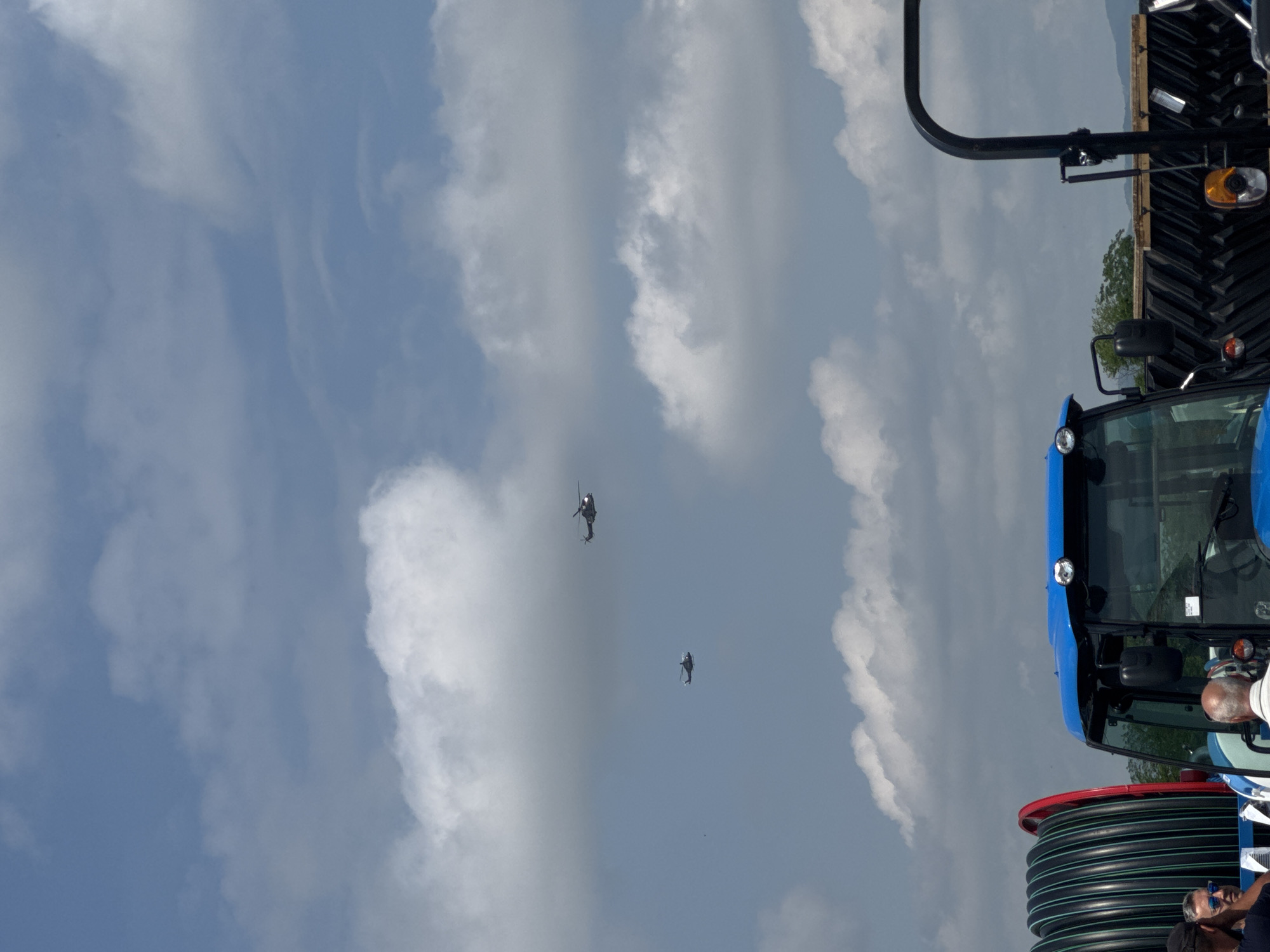 The image, oriented correctly, captures an outdoor scene under a bright, partly cloudy sky, likely during midday or early afternoon. The sky, a mix of blue and scattered white clouds, occupies most of the frame.
Two individuals are visible flying personal aerial devices in the mid-ground. These devices resemble small, open-cockpit helicopters or advanced jetpacks, each with a person piloting it. One flyer is positioned higher and further away, while the other is lower and closer to the viewer, both silhouetted against the sky.
In the bottom right foreground, the front section of a blue tractor, likely a New Holland model, is prominent. Features like its cabin, a headlight, and a large black roll bar extending upwards are visible. Adjacent to the tractor, a large, black industrial spool of coiled tubing or hose is also present.
Several people are partially visible in the bottom right, near the tractor and the spooled tubing. One man with a bald head and a white t-shirt is clearly looking upwards, seemingly observing the flying devices. Another person wearing sunglasses is also present.
The scene appears to be an outdoor event or demonstration, possibly combining an air display with an agricultural or machinery exhibit, set against a backdrop of distant hills and scattered trees. The location is Madzherito, Bulgaria. No discernible text is visible on any of the subjects.